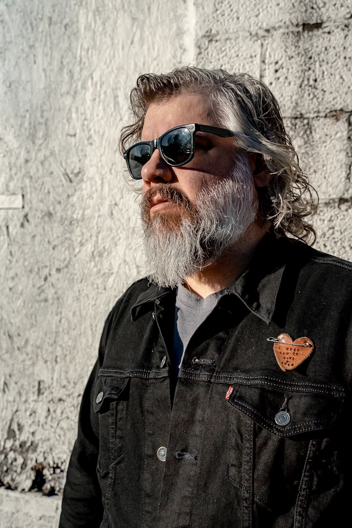 Man with sunglasses, gray beard, and long hair stands against a textured wall wearing a black jacket with a heart-shaped pin.