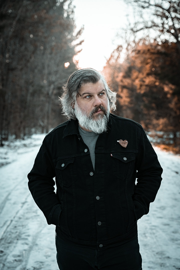 Man with gray hair and beard wearing a black jacket outdoors in a snowy forest during sunset.