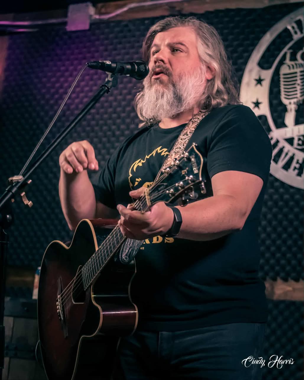 Man with long gray hair and beard singing into a microphone while playing an acoustic guitar.