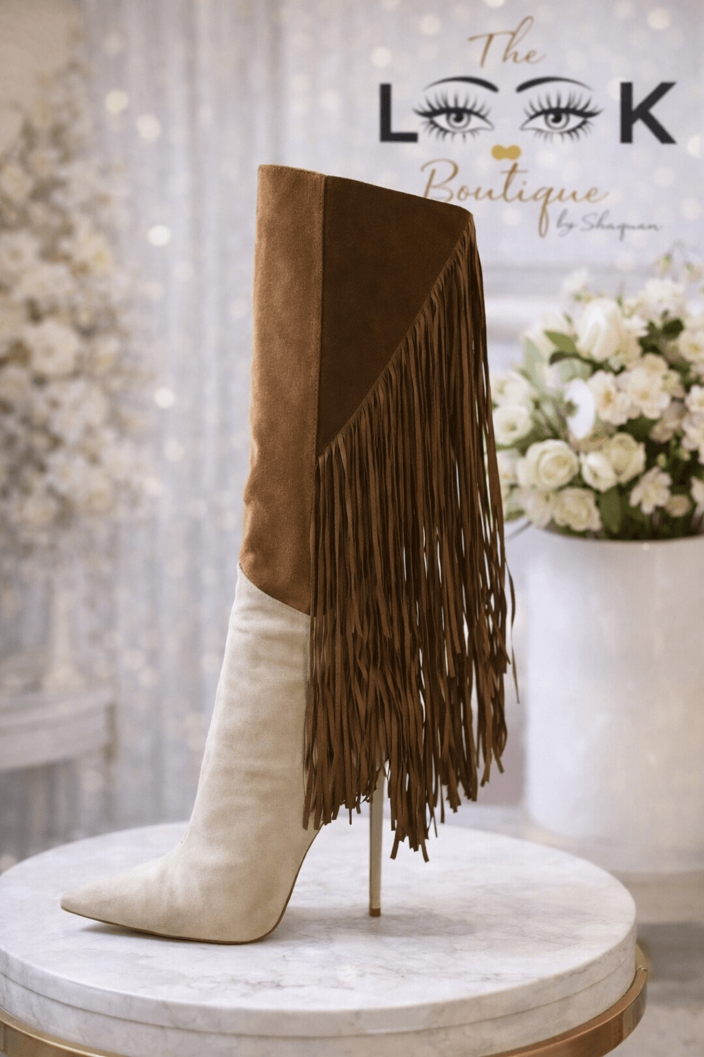 High-heeled boot with fringe detail, placed on a round marble surface, with a floral arrangement in the background.