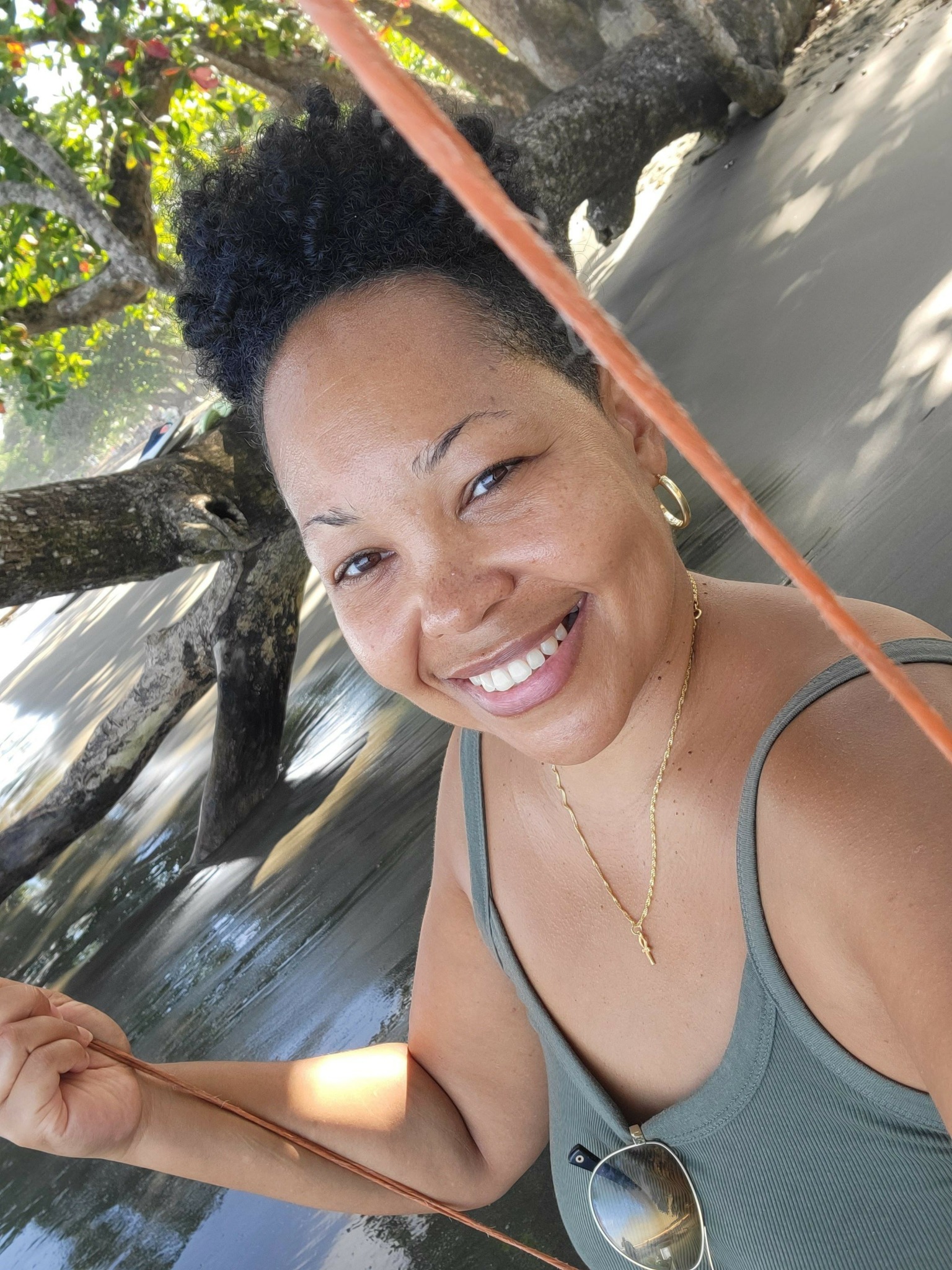 Smiling woman with short curly hair holding a swing, outdoors near a tree and water.