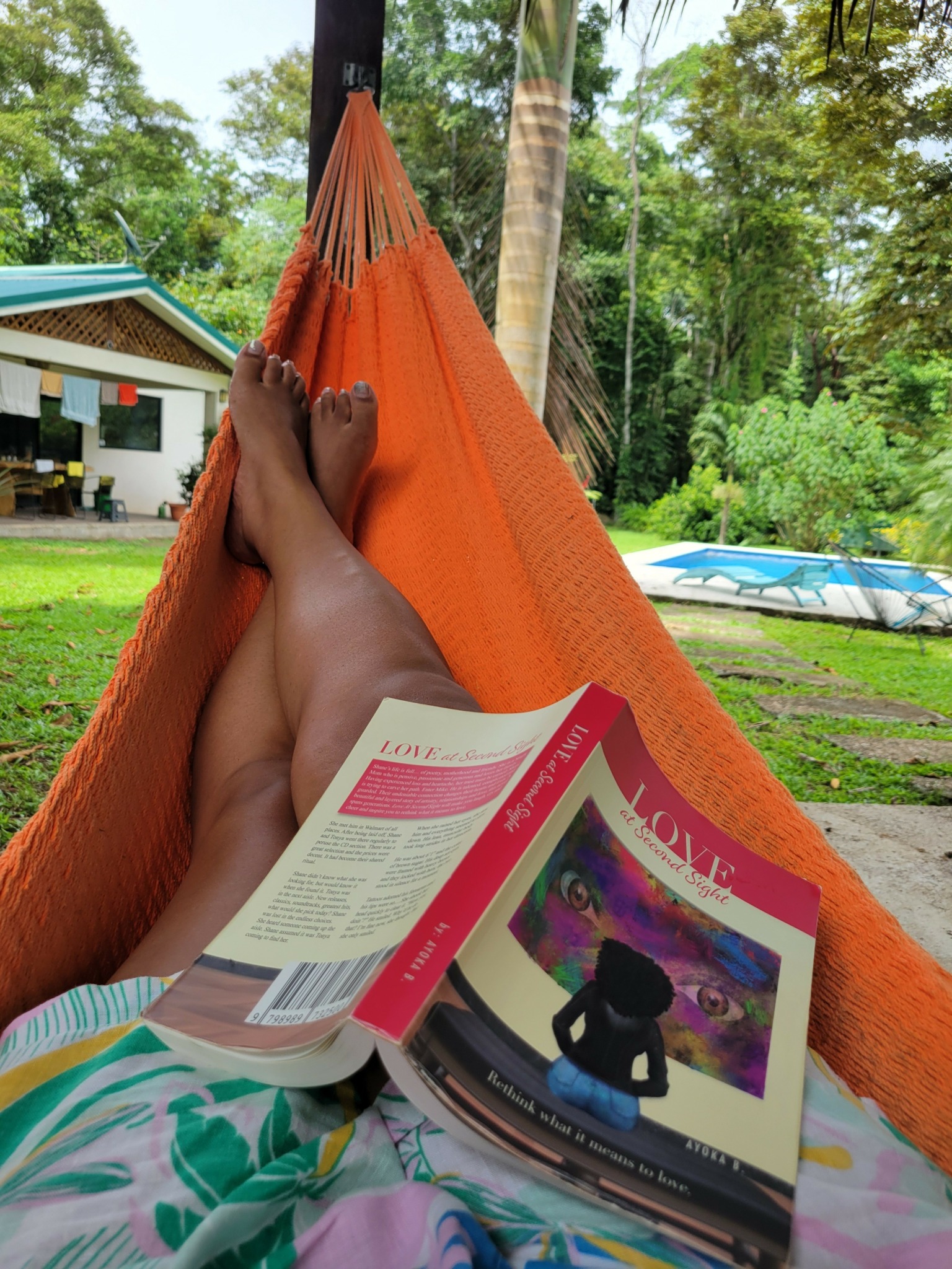 Person relaxing in hammock outdoors, reading a magazine with a colorful cover, surrounded by trees and greenery.