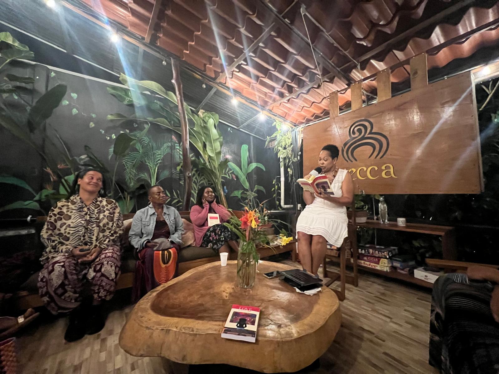 Four women sitting at a table in a tropical-themed cafe, one woman standing reading a book, large sign with cafe logo behind.
