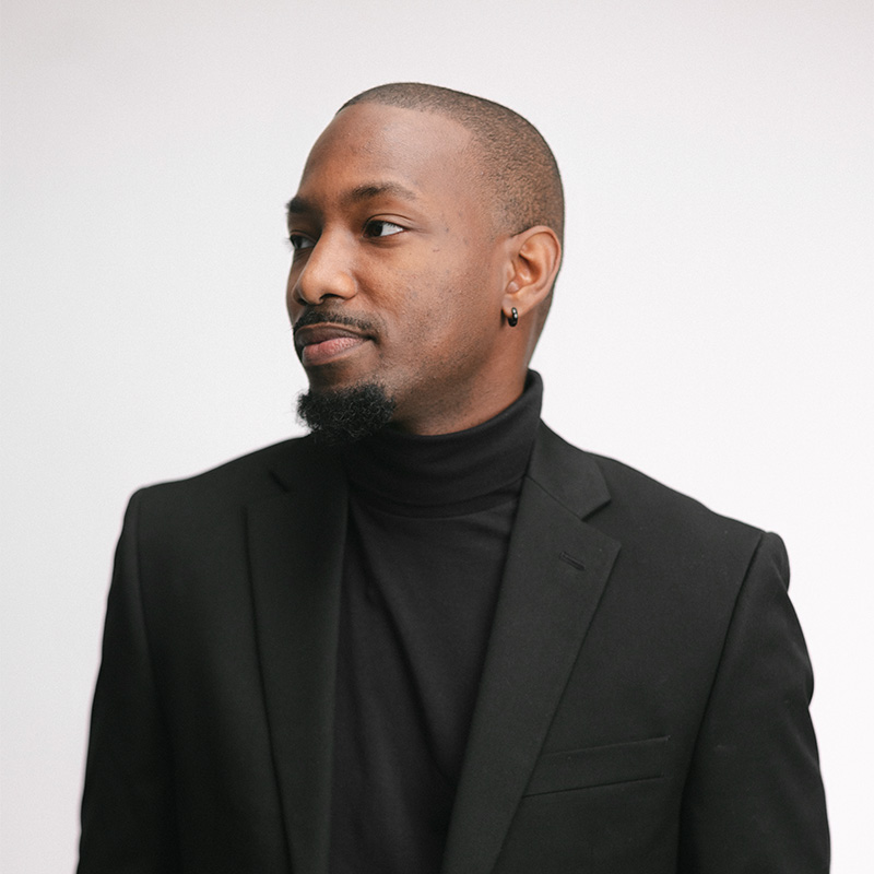Man with short hair, beard, wearing black turtleneck and blazer, looking to the side against a plain background.