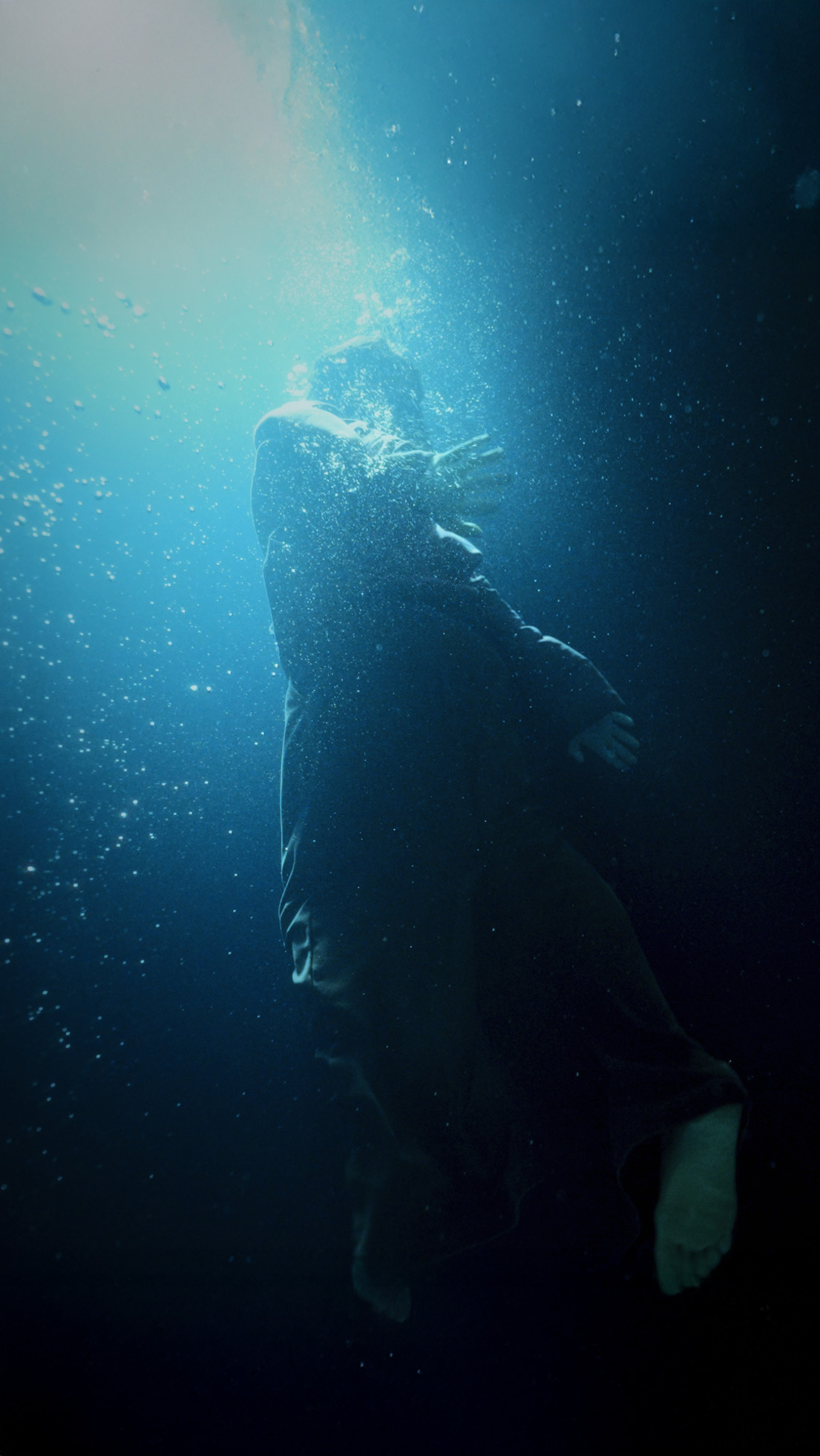 Person underwater, surrounded by bubbles, with light filtering from above, wearing dark clothing.