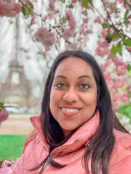 Smiling woman with long dark hair outdoors among pink flowering trees.