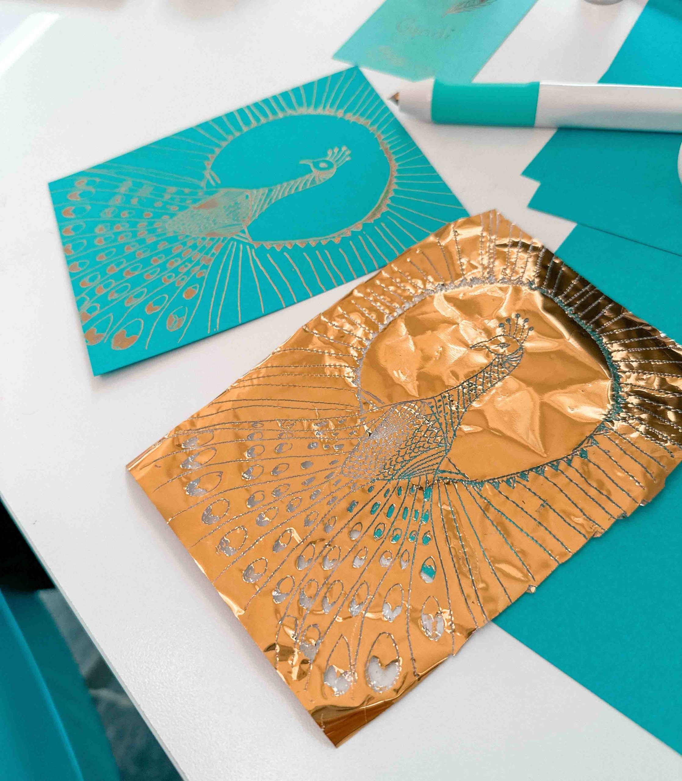 Two decorated cards with peacock designs, one in blue and gold foil, on a white surface with markers and paper.