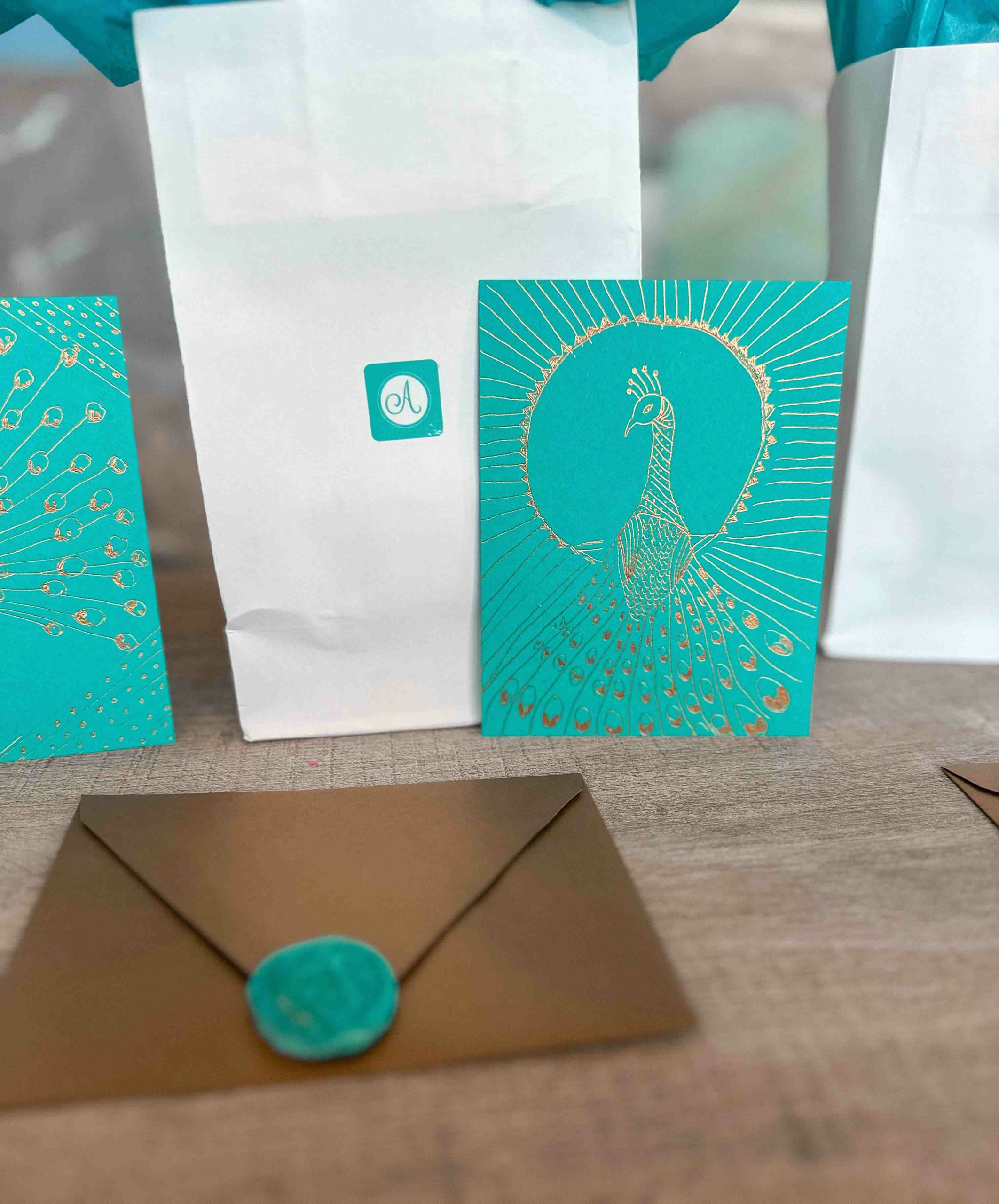 White gift bags with teal designs and a teal greeting card with a peacock illustration, on a wooden surface.