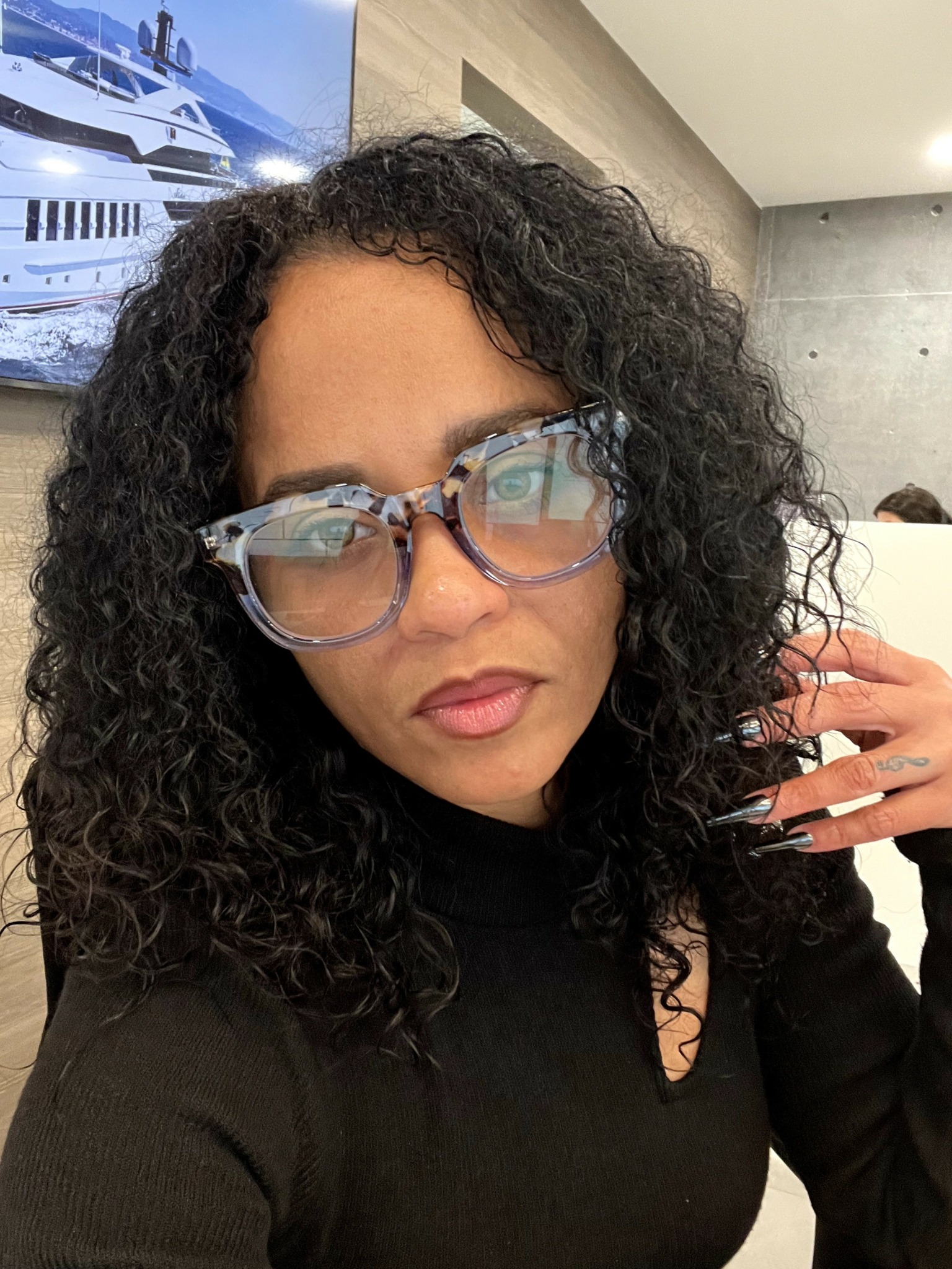 Woman with curly hair wearing large patterned glasses and black top, indoors with a yacht picture in background.
