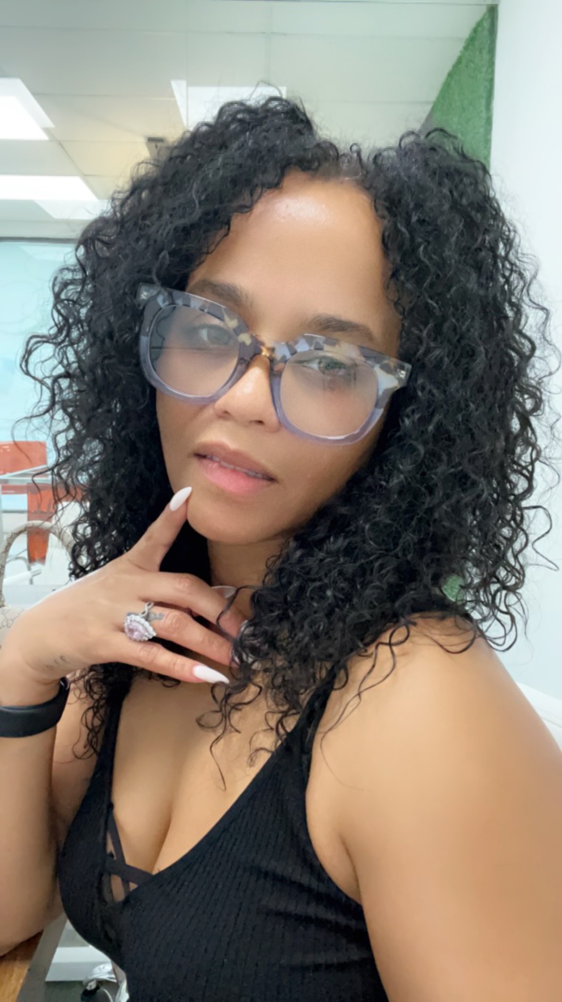 Woman with curly hair wearing glasses and a black top, touching her chin, indoors.