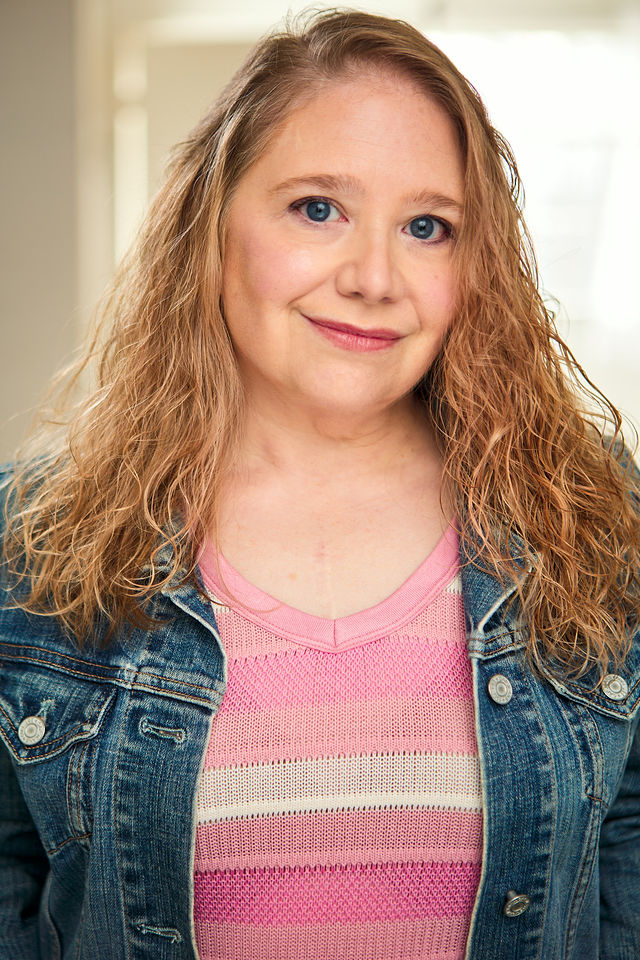 Young woman with curly blonde hair smiling, wearing a pink and gray striped shirt and denim jacket indoors.