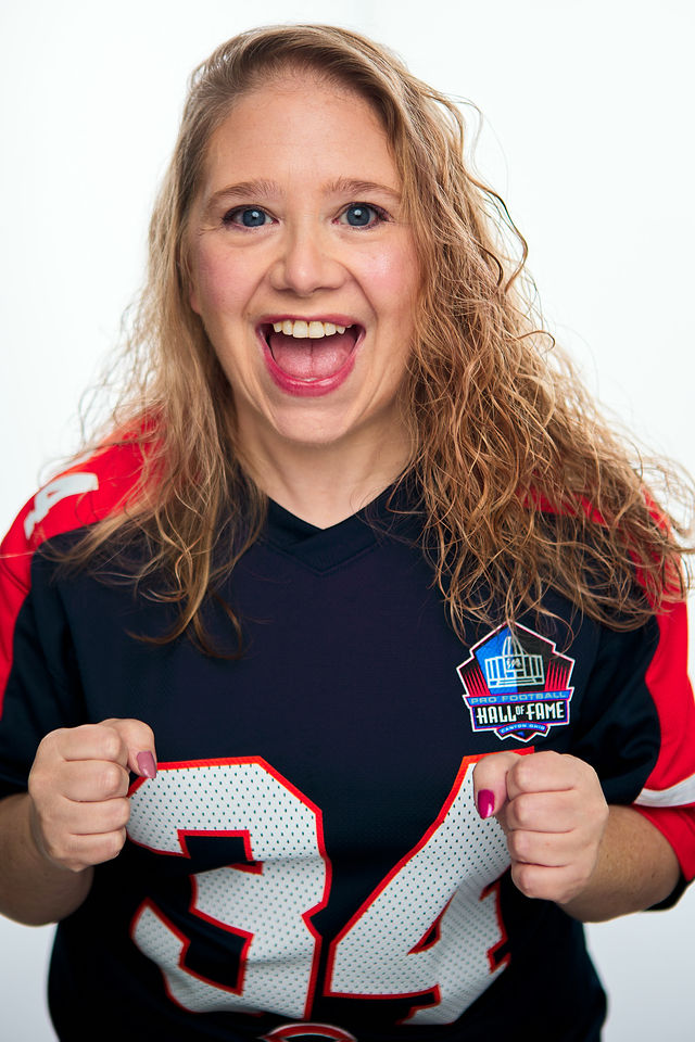 Young woman with curly hair smiling, wearing a sports jersey with a Hall of Fame patch, holding her fists up.