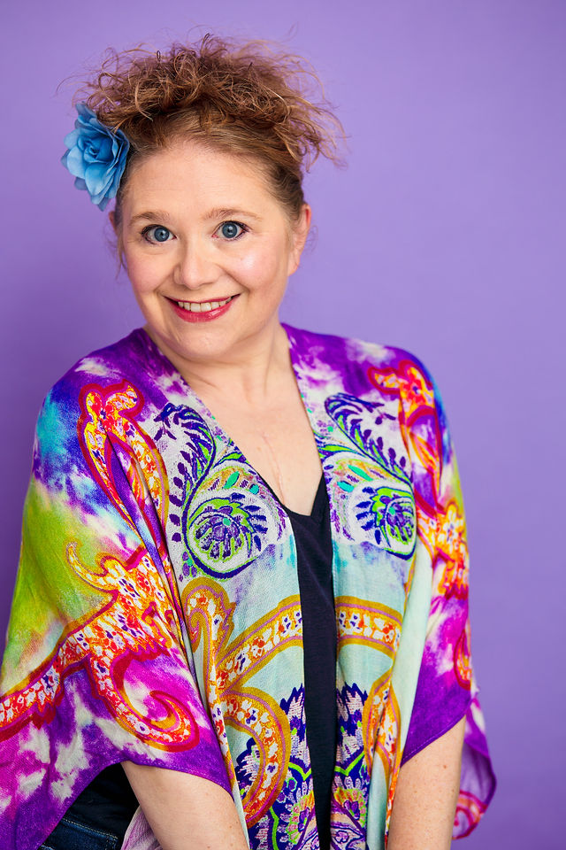 Woman with curly hair and a blue flower hair accessory smiling, wearing a colorful patterned jacket against purple background.