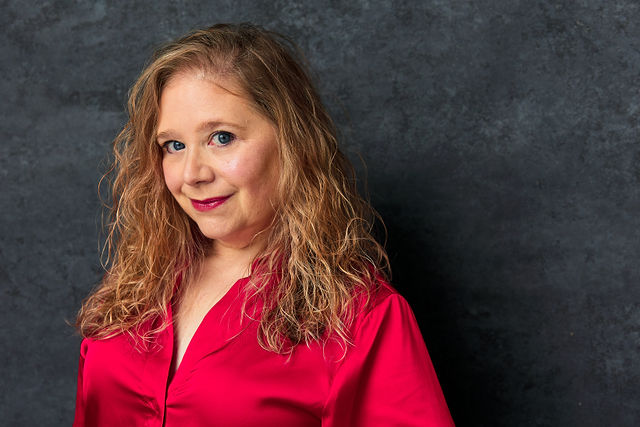 Woman with curly blonde hair wearing a red blouse, standing against a dark textured background.