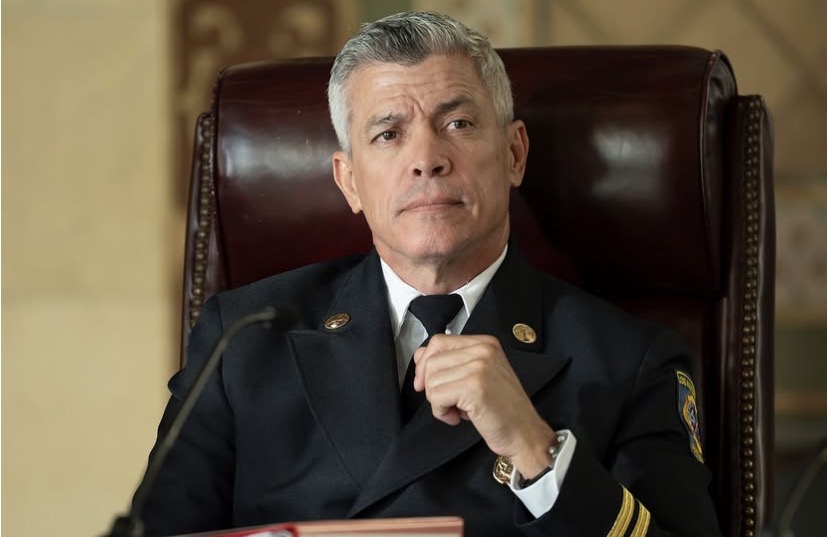 Older man in a uniform sitting in a leather chair, looking serious, with a hand near his chin.