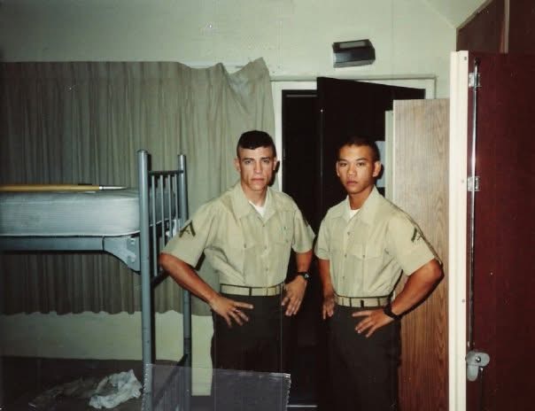 Two young men in uniform standing in a room with a bunk bed and a wooden partition.