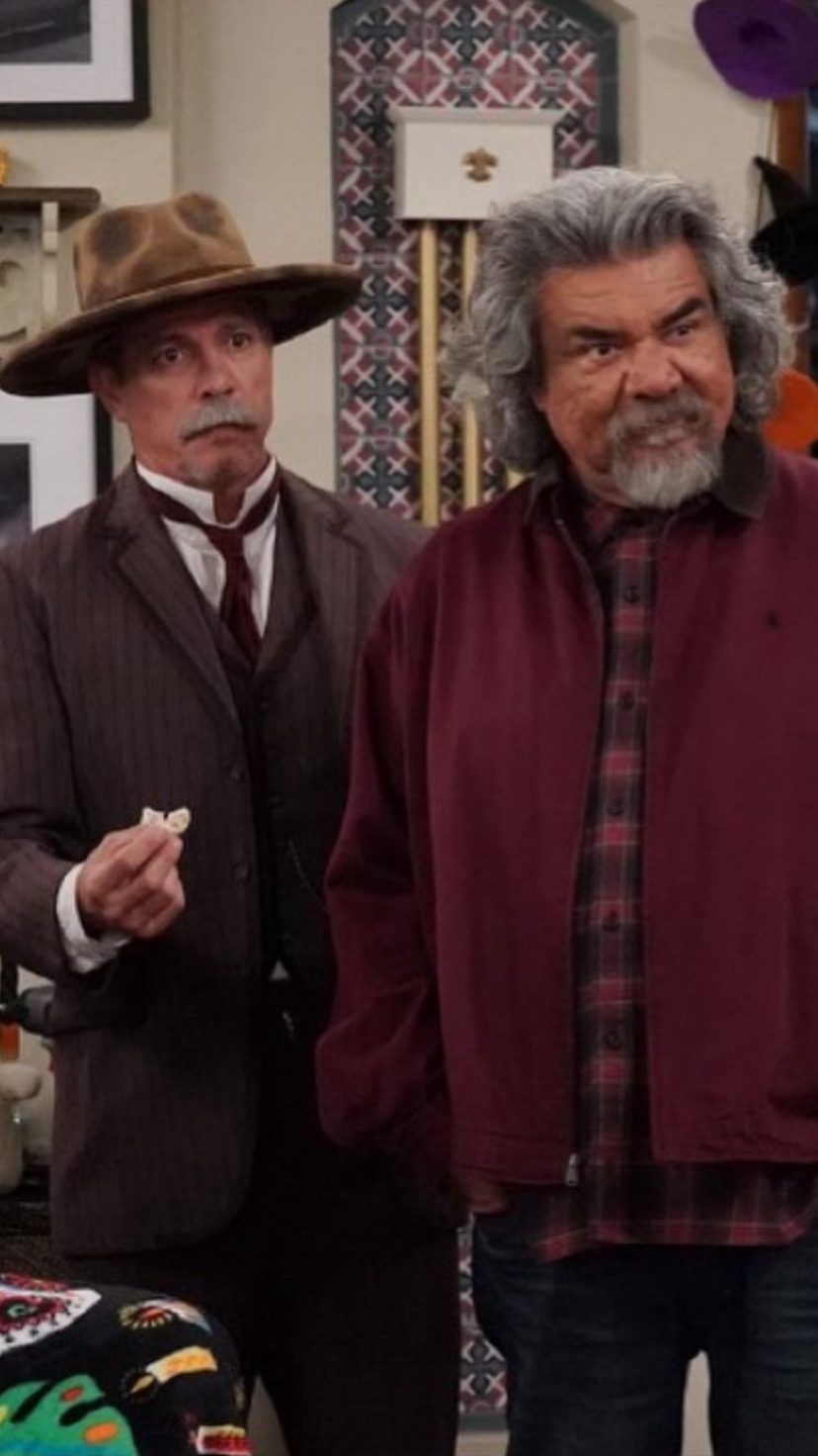 Two men standing indoors, one in a suit and hat, the other in a maroon jacket with gray hair and beard.