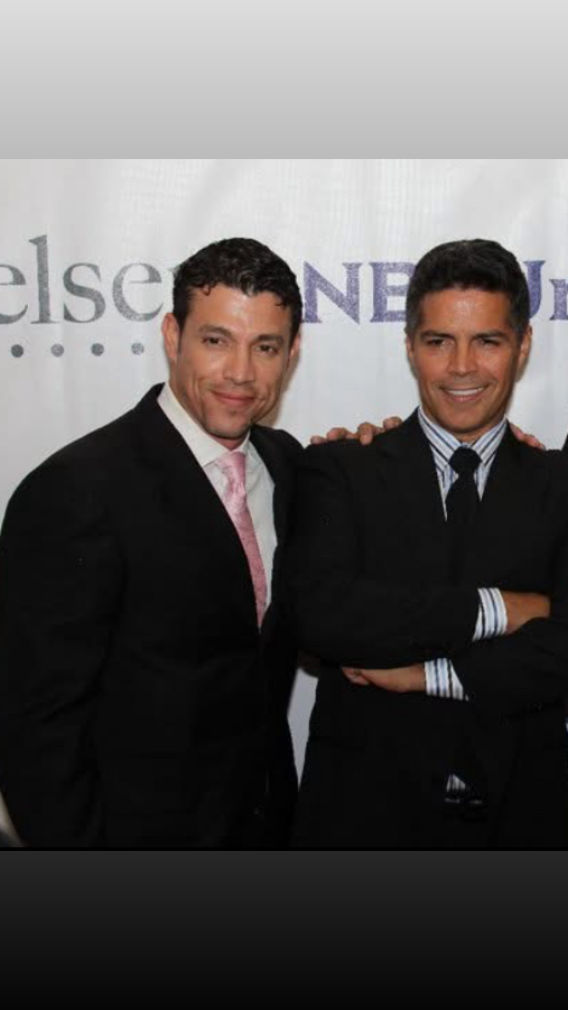 Two men in suits standing side by side, smiling, with a blurred background featuring text.