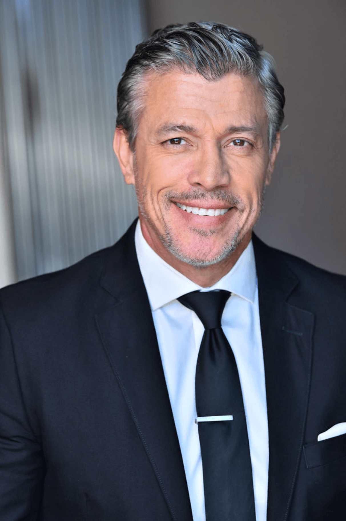 Man in suit smiling, wearing a white shirt and black tie, with gray hair and beard, standing indoors.