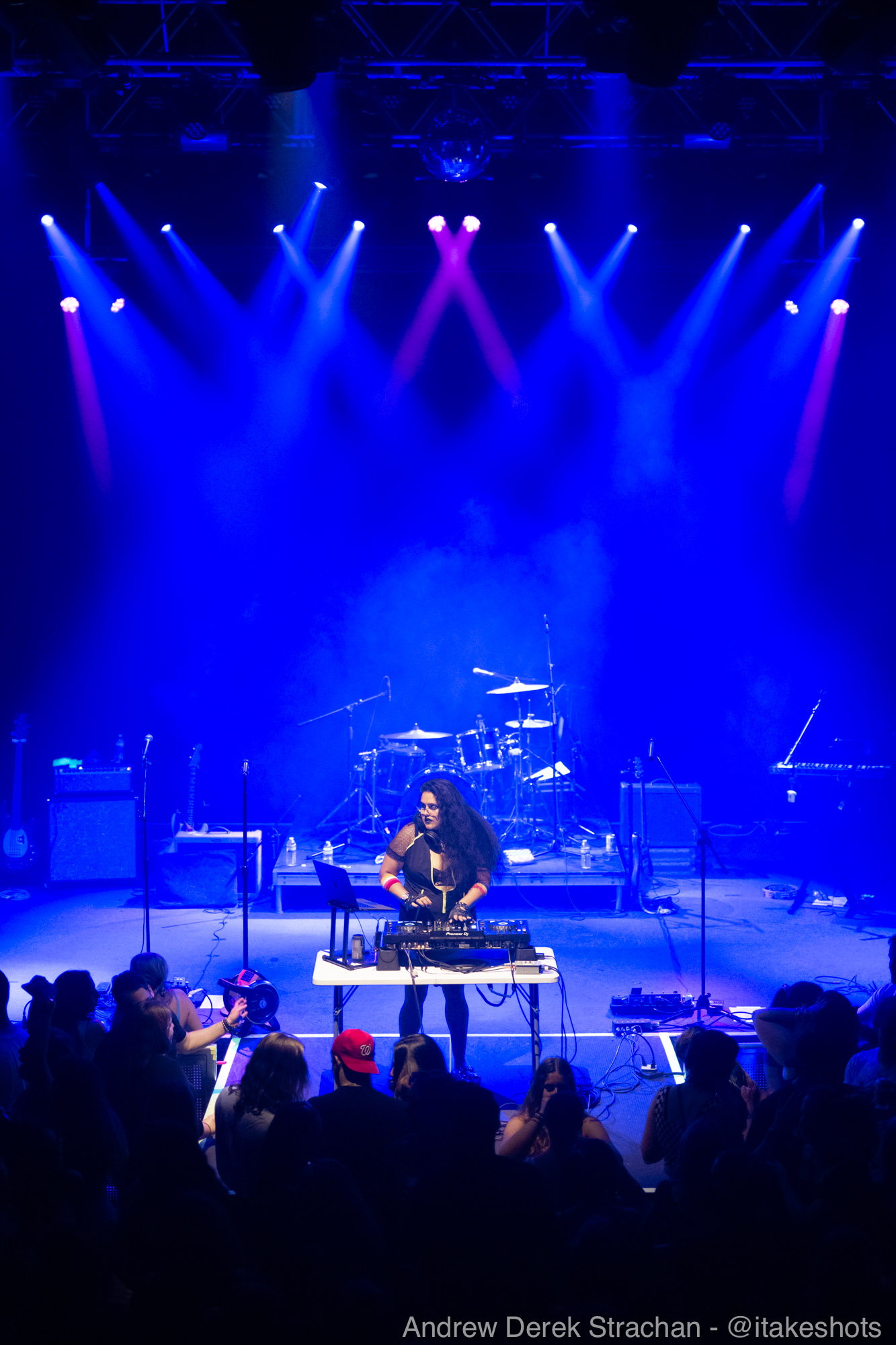 Performer on stage with musical instruments, colorful stage lights, audience in foreground, concert setting.