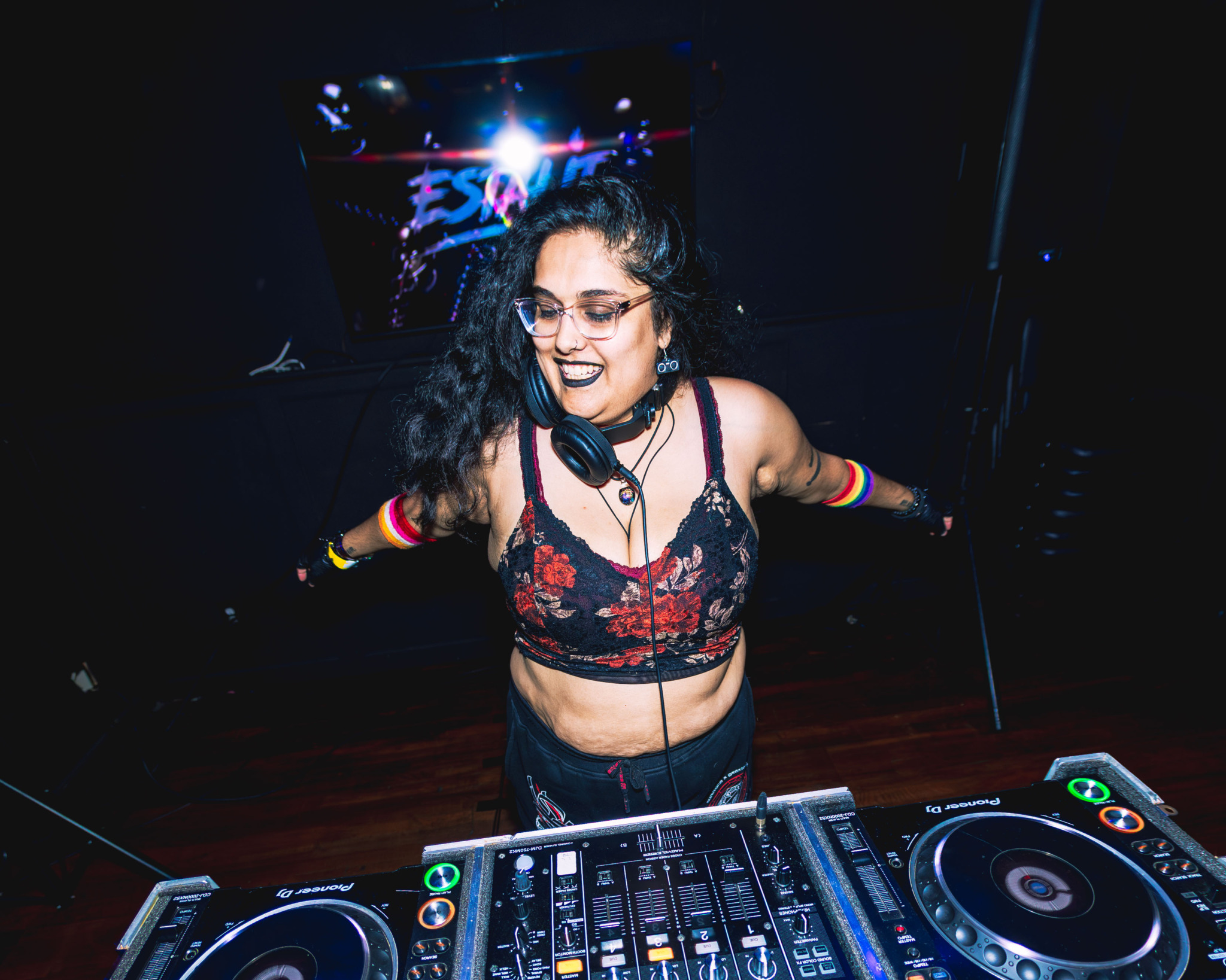 Female DJ with curly hair and glasses, wearing a floral crop top, standing behind DJ equipment in a dark club.