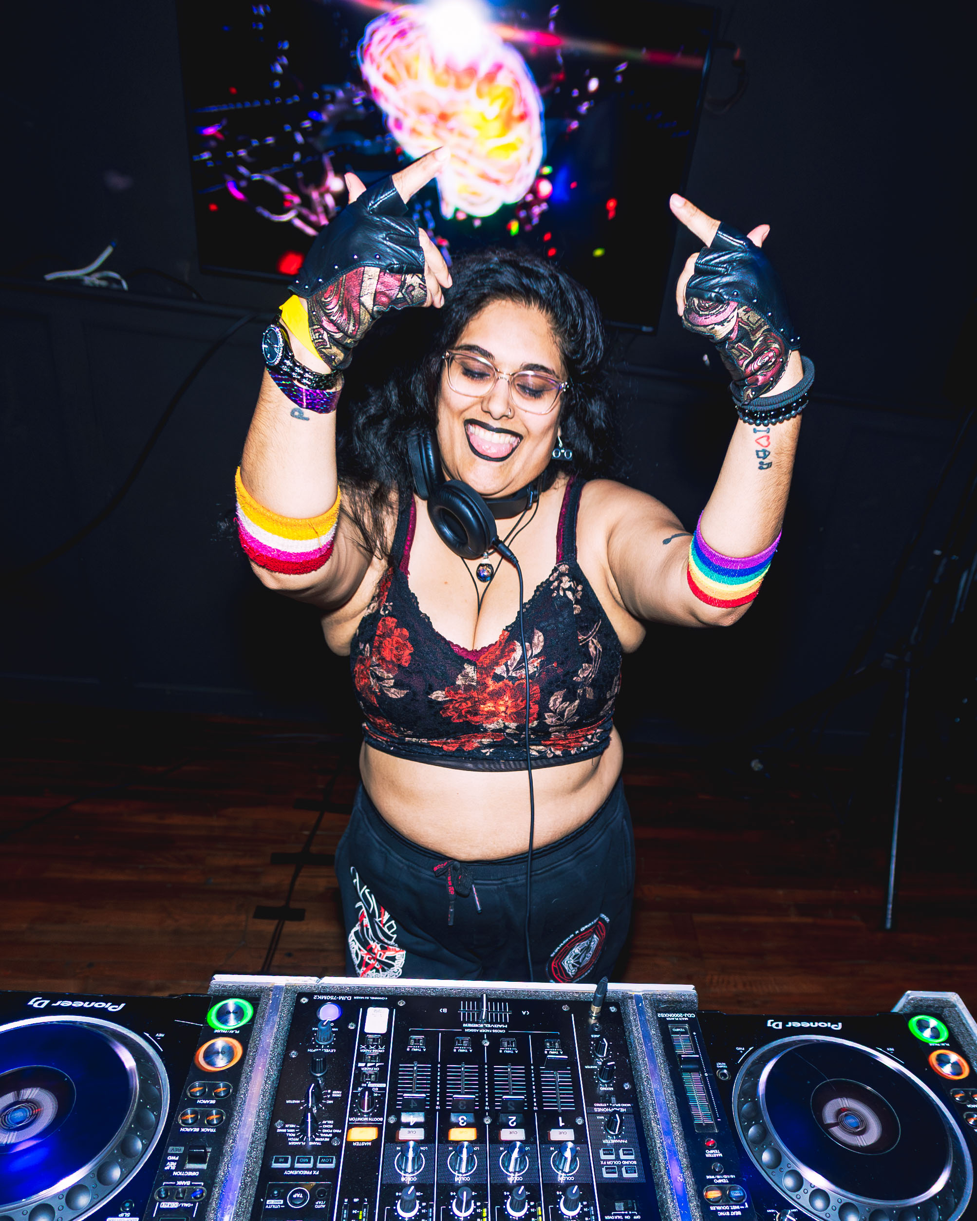 DJ woman with headphones, colorful arm bands, and gloves, smiling with hands raised in a club setting.