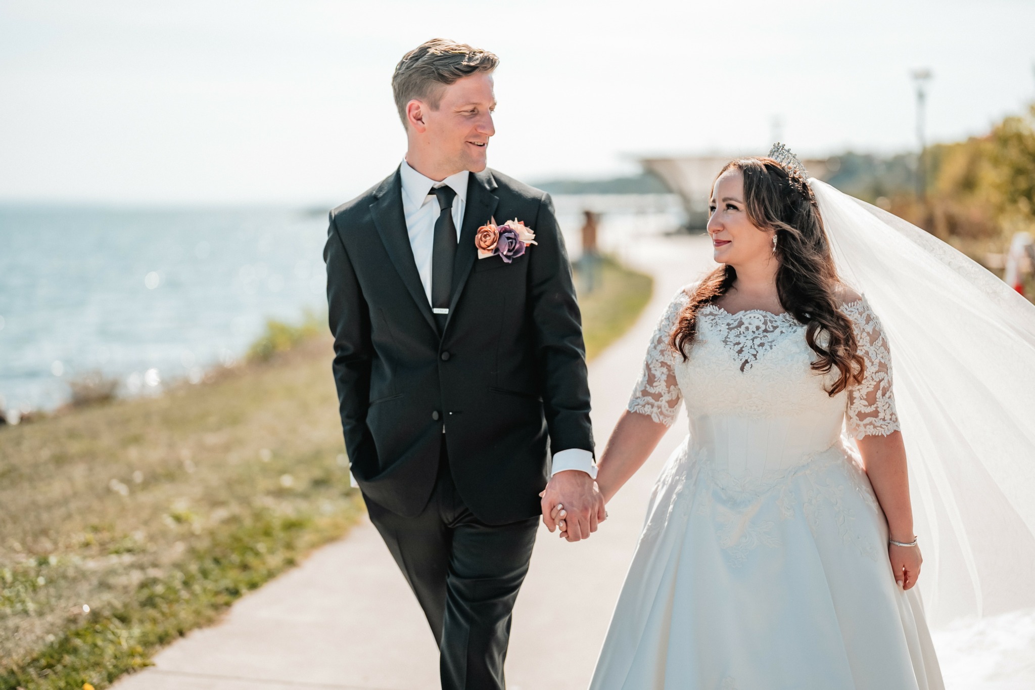 A bride and groom holding hands outdoors near water, smiling at each other, with a cloudy sky and shoreline in background.