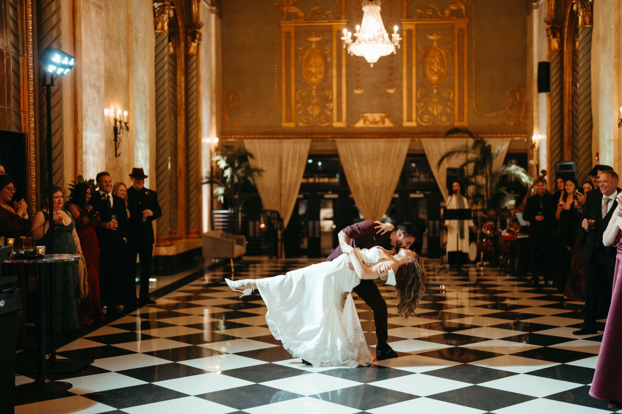 Couple dancing in a grand ballroom with checkered floor, surrounded by guests and ornate decor.
