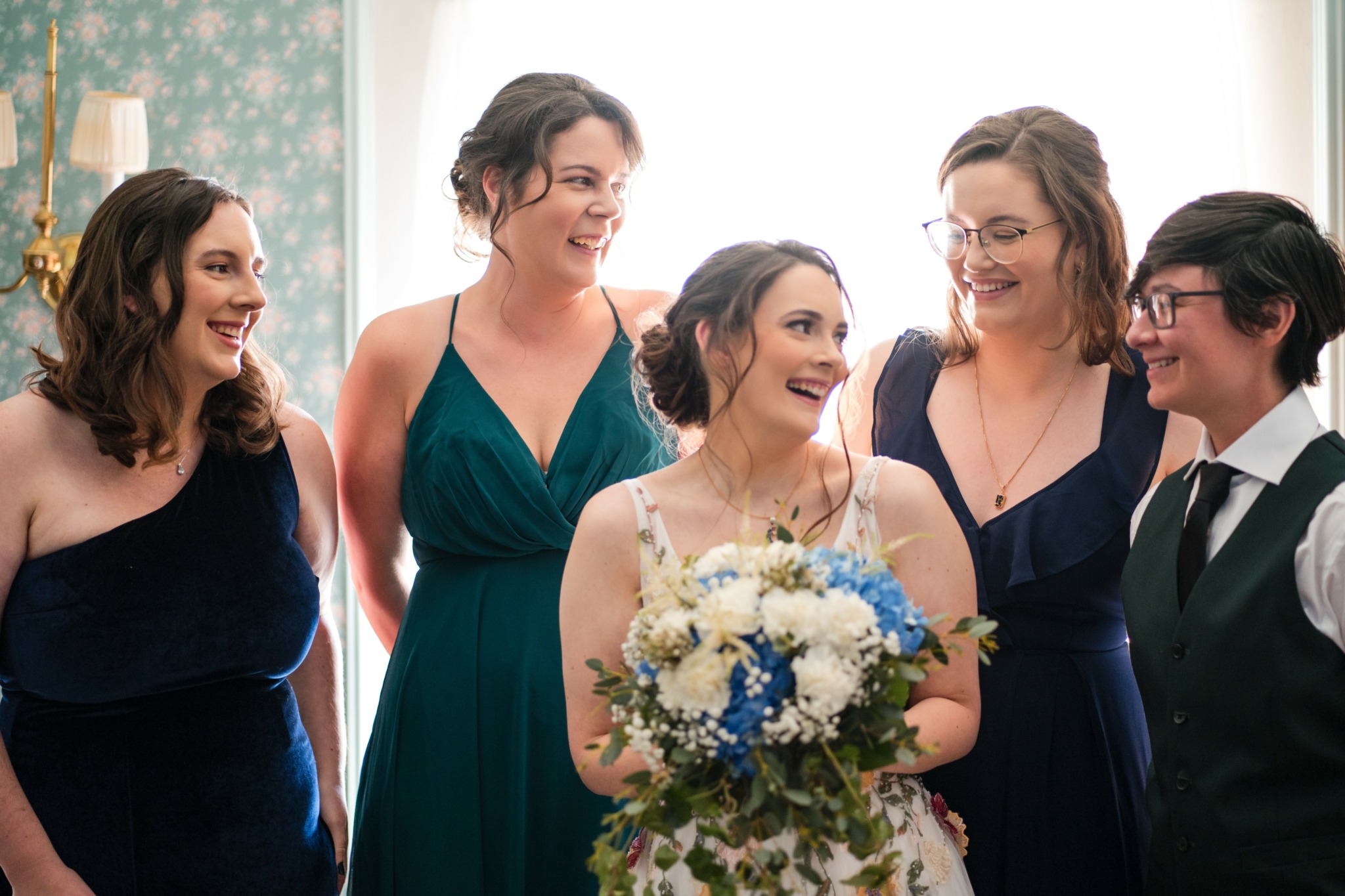 Group of five women smiling, one holding a bouquet of white and blue flowers, indoors with light background.