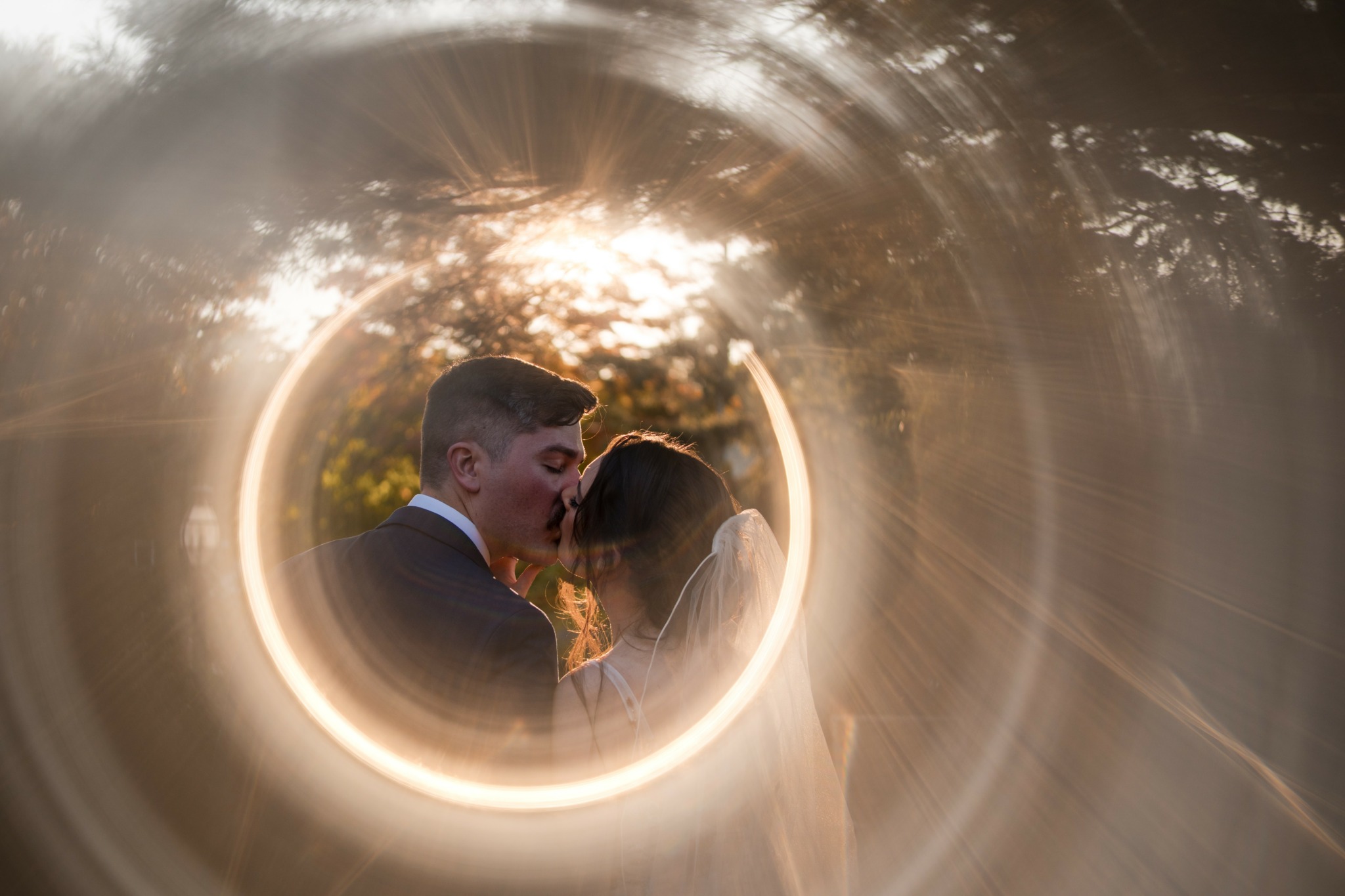 Couple facing each other outdoors with sunlight behind trees, seen through a circular light or lens effect.