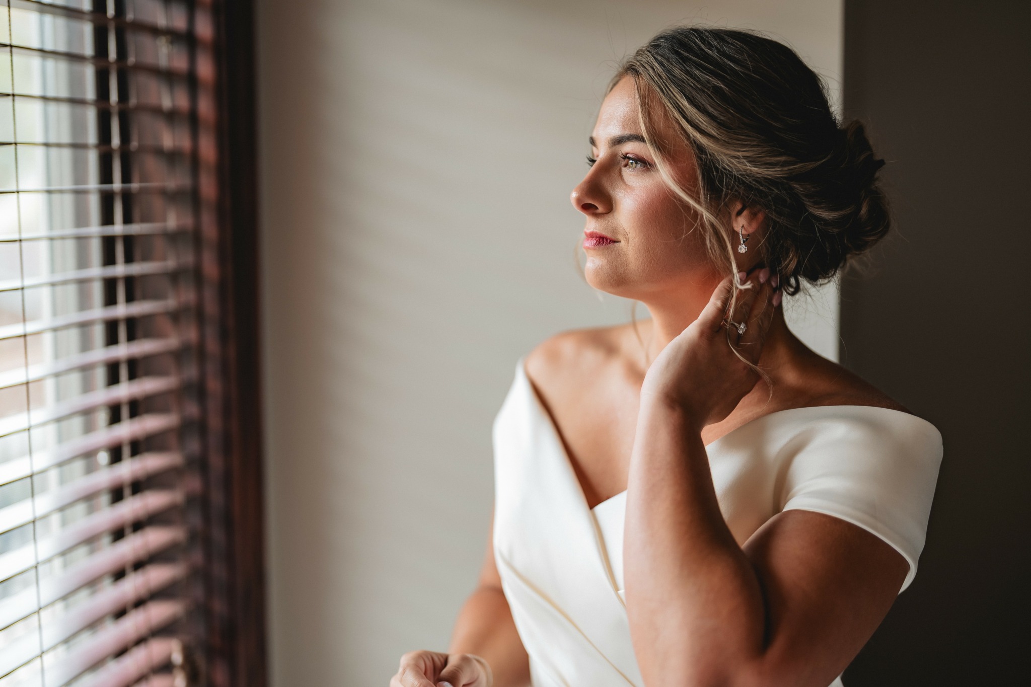 Woman with styled hair wearing a white dress looking out a window with blinds.