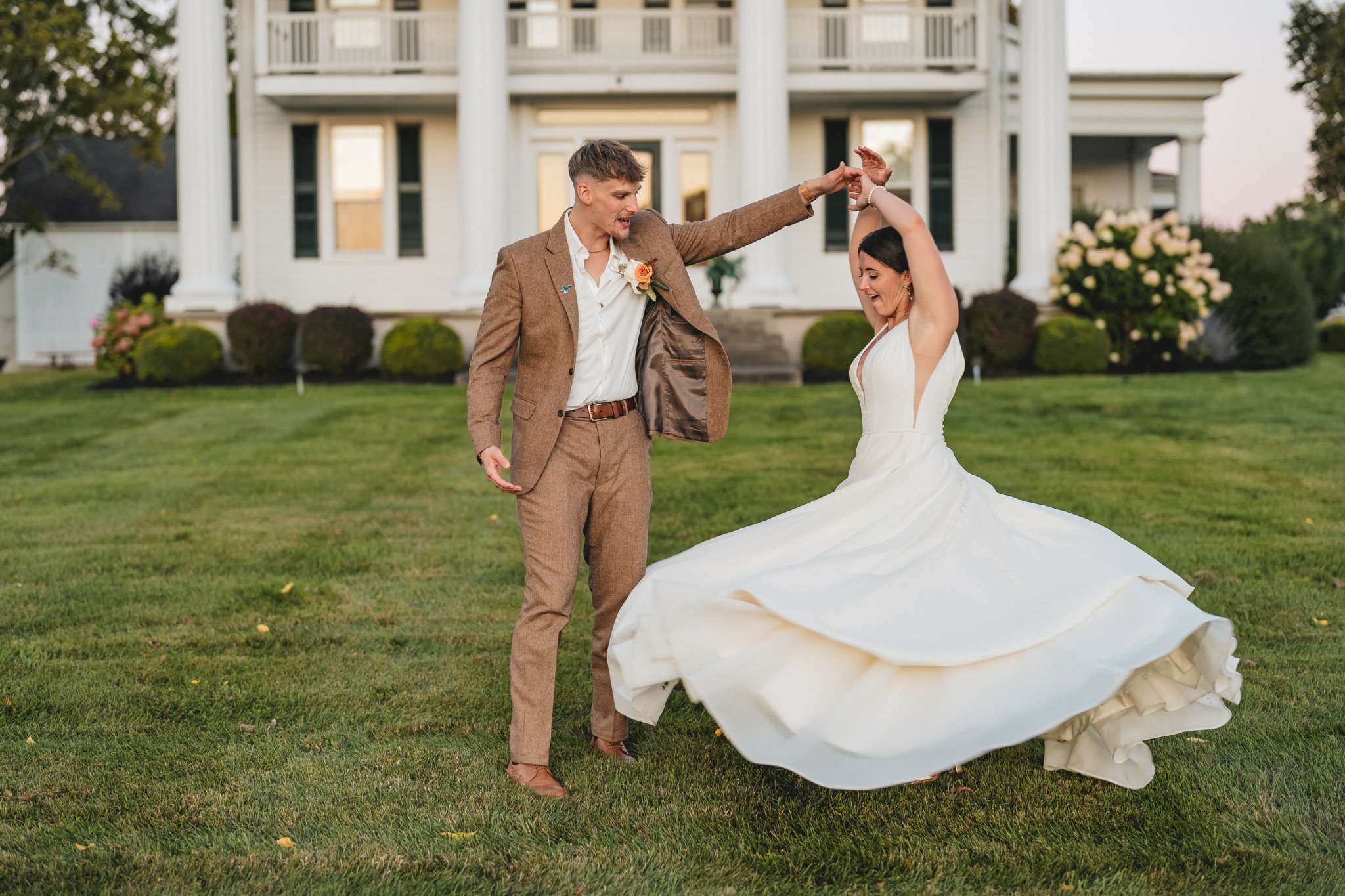 Bride twirling in a white wedding dress, groom in brown suit, on a lawn in front of a large white building.