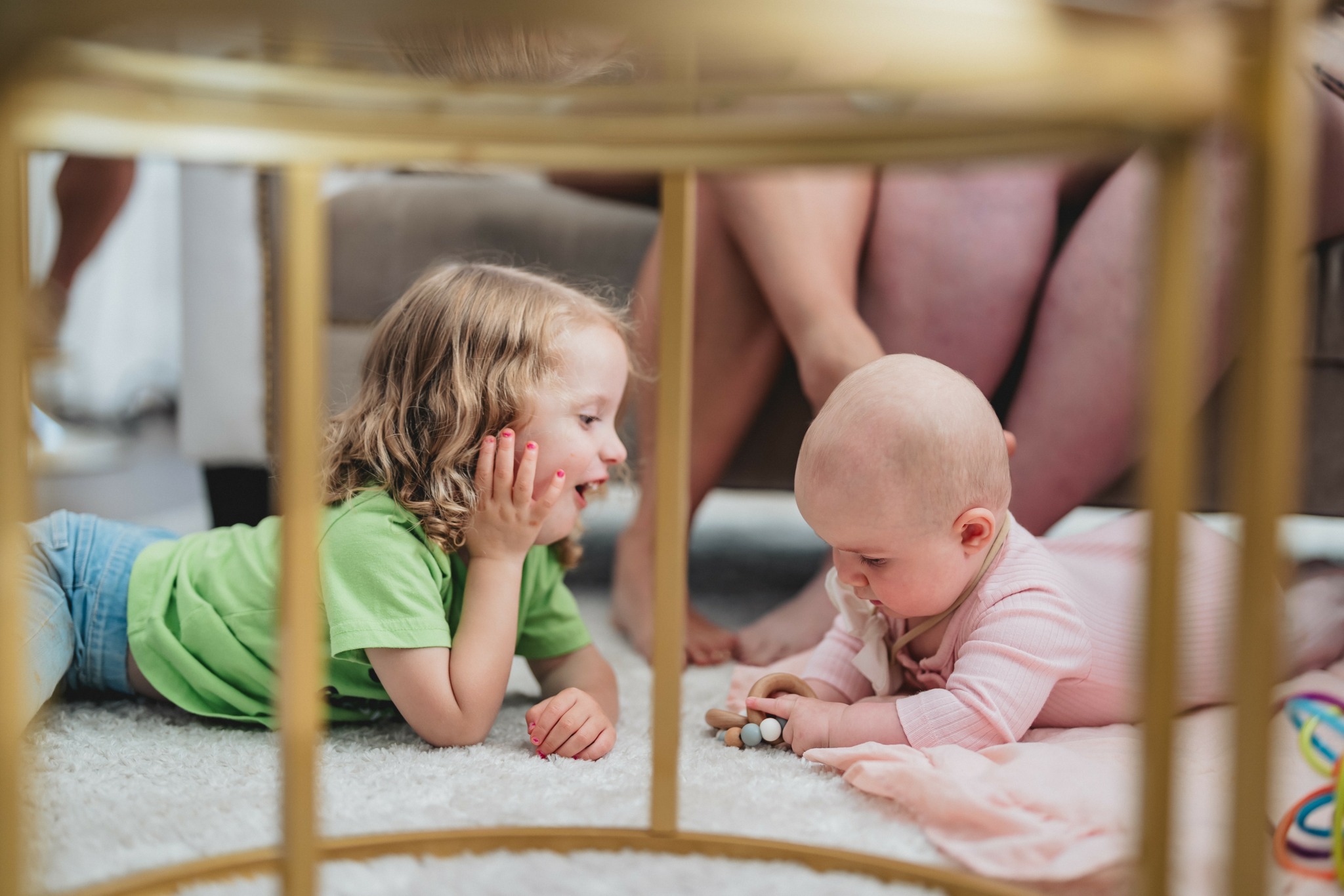 Two children lying on the floor, facing each other, with a person sitting nearby. The girl has blonde hair and a green shirt.