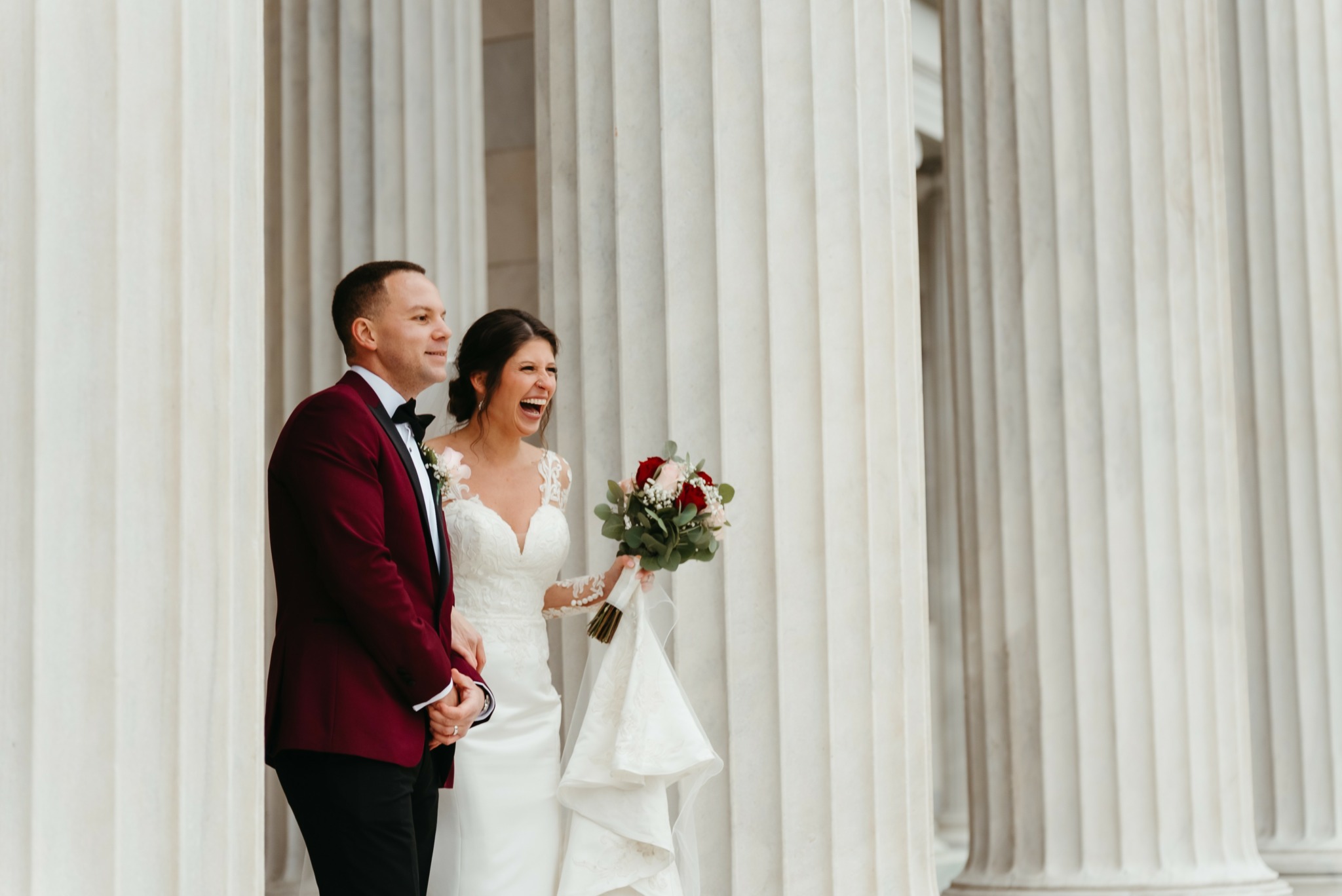 Bride and groom standing between large columns, smiling, with the bride holding a bouquet of flowers.