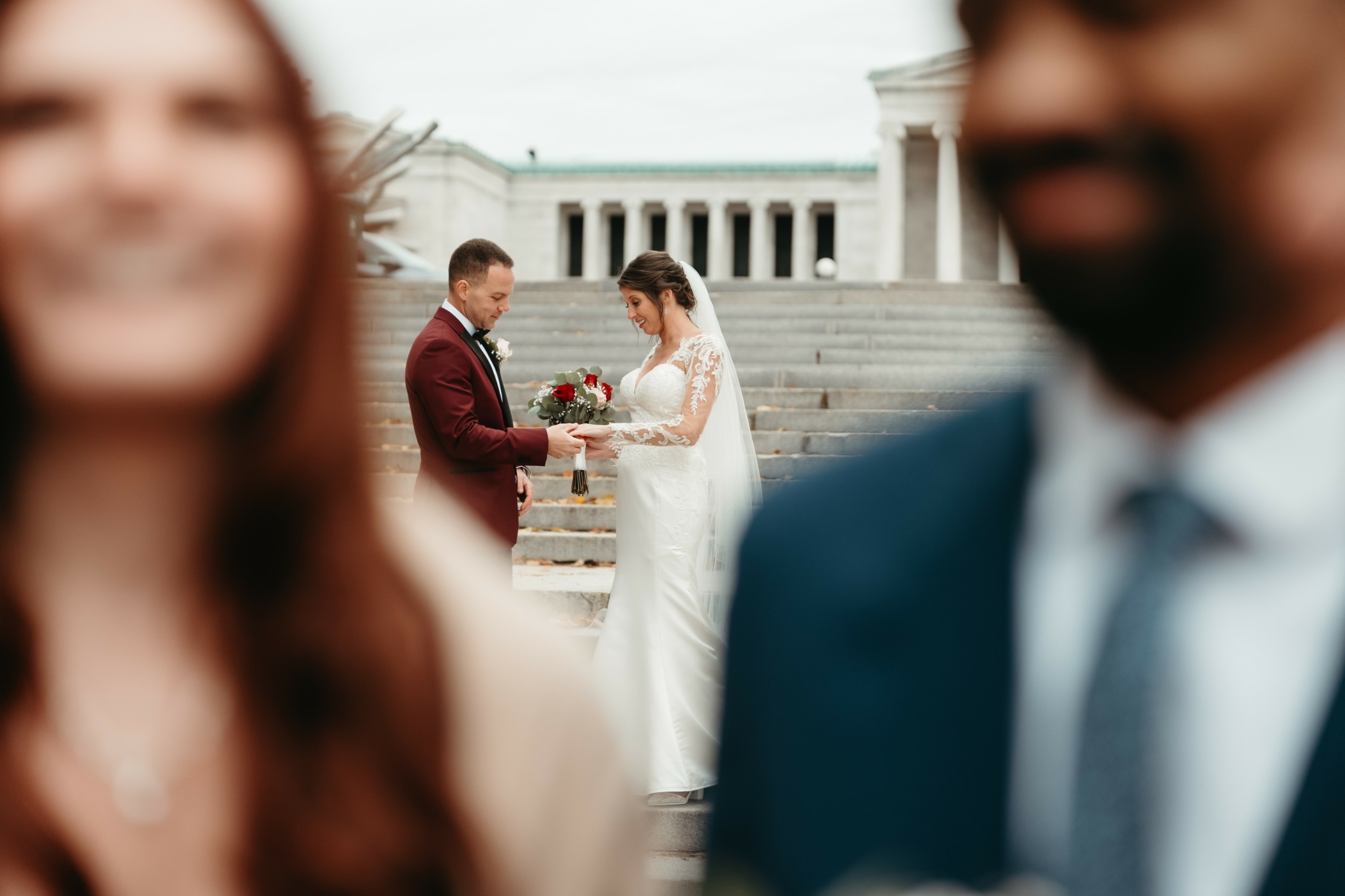 Bride and groom exchanging rings on steps, with blurred woman and man in foreground, building in background.