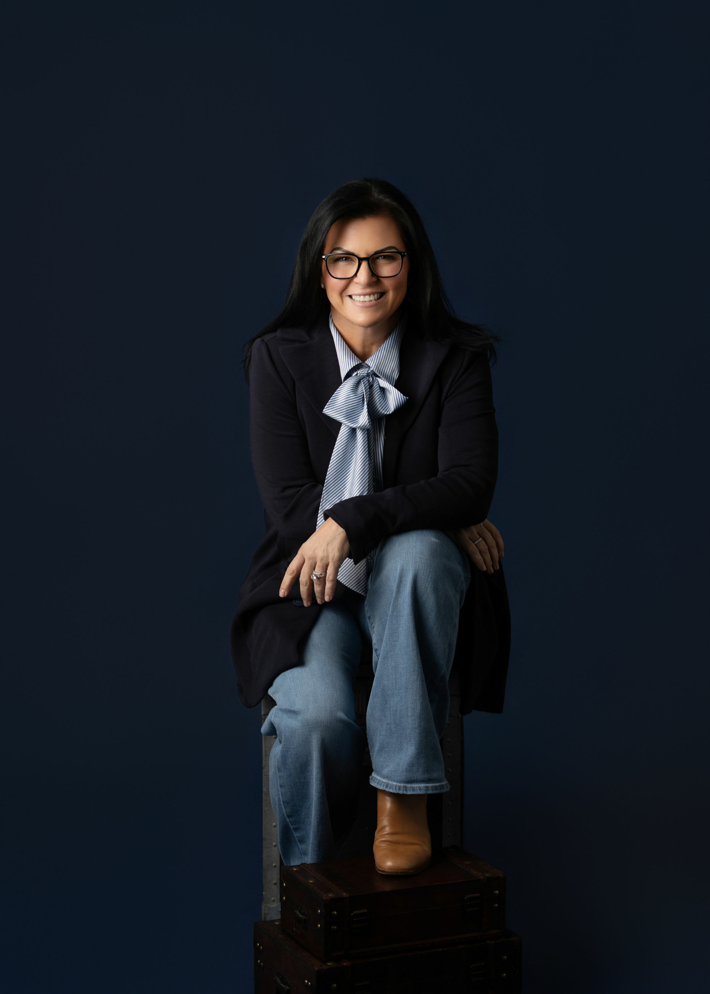 Woman with glasses sitting on a box, smiling, wearing a dark blazer, light blue shirt, jeans, and brown boots.