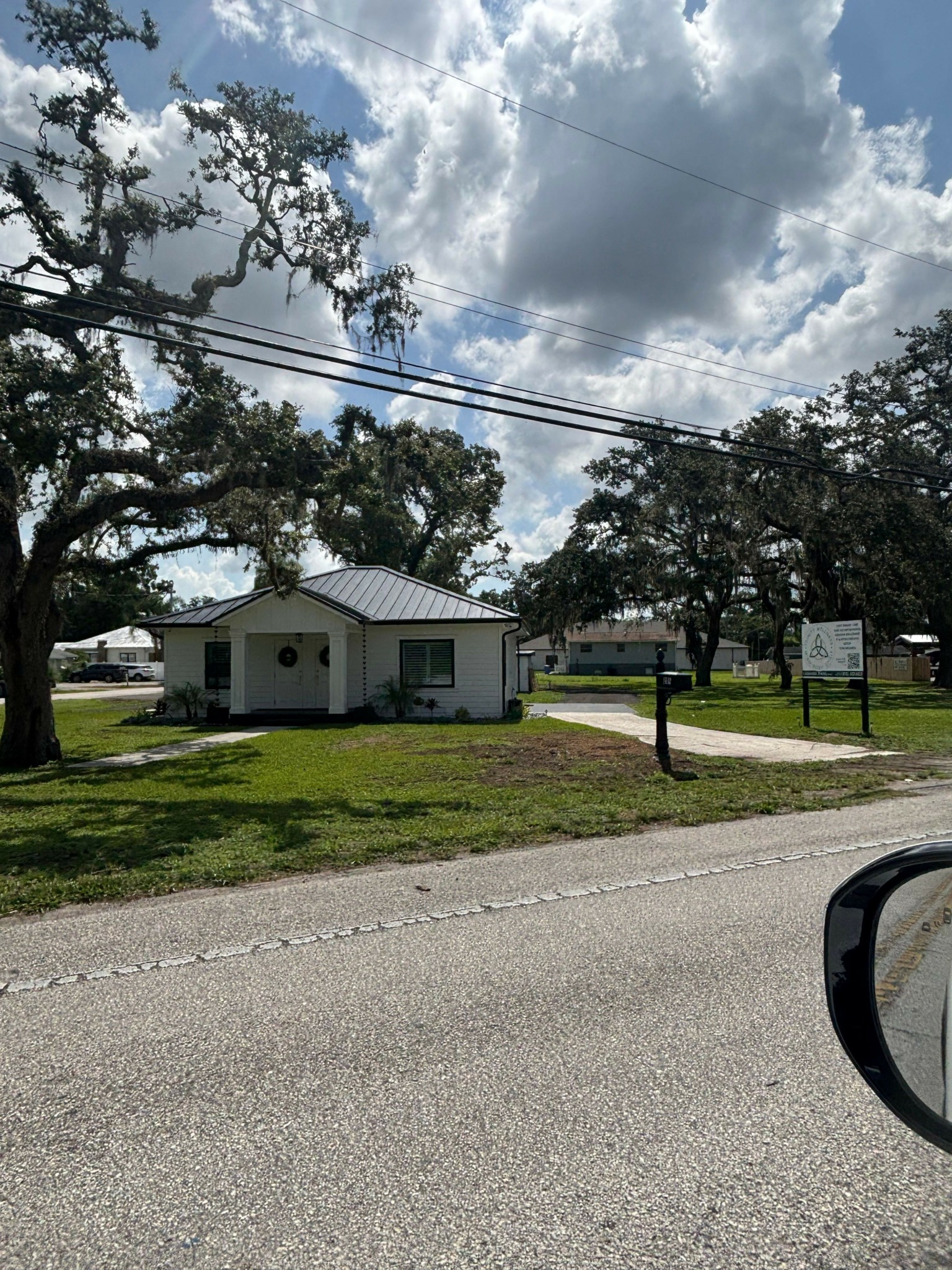 Small building with a metal roof, trees, and a sign on a grassy area beside a road under a partly cloudy sky.