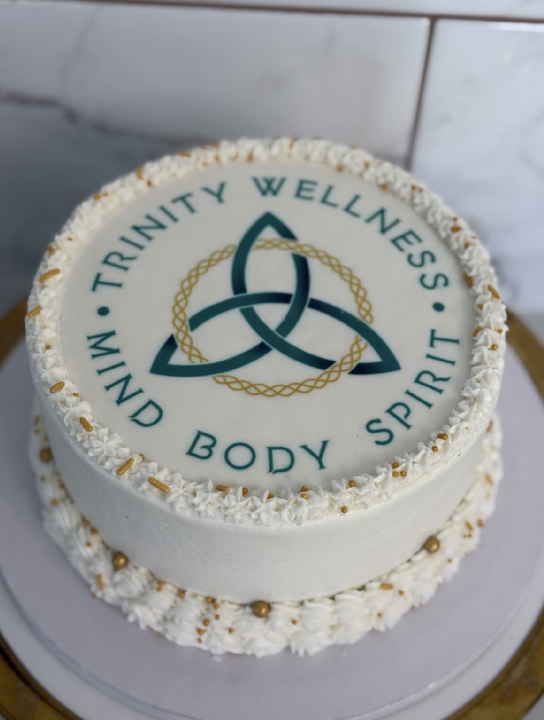Round cake with white icing, logo with triquetra symbol, and words 'Trinity Wellness' and 'Mind Body Spirit'.
