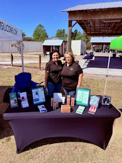 Two women standing behind a table with various products outdoors, under a pavilion, with a fence and trees in background.