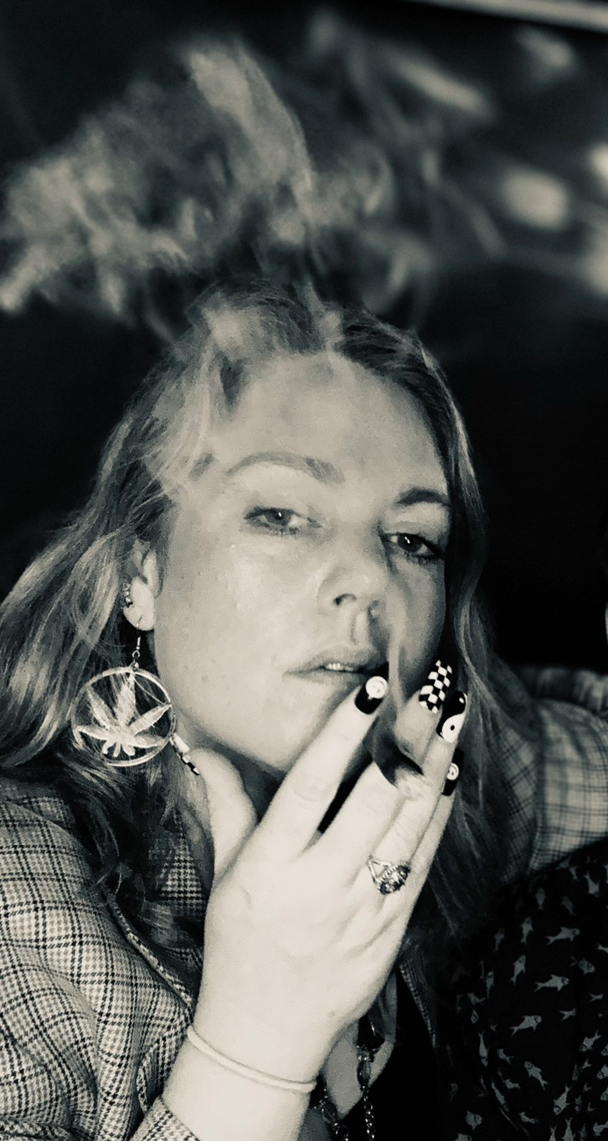Young woman with long hair, wearing earrings, holding fingers near lips, in a dark setting with smoke or vapor.