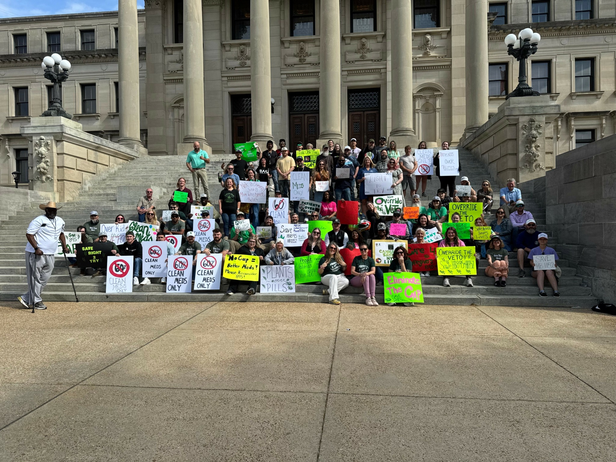 Group of people protesting on steps of a large government building, holding signs and banners.
