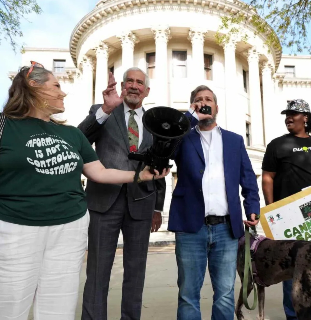 Group of four people standing outside a government building, one holding a megaphone, others smiling and talking.