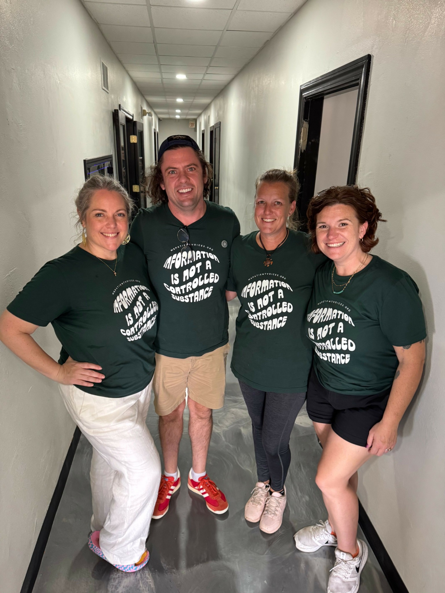 Four people standing in a hallway, smiling, wearing matching dark green T-shirts, with white text, and casual pants or shorts.