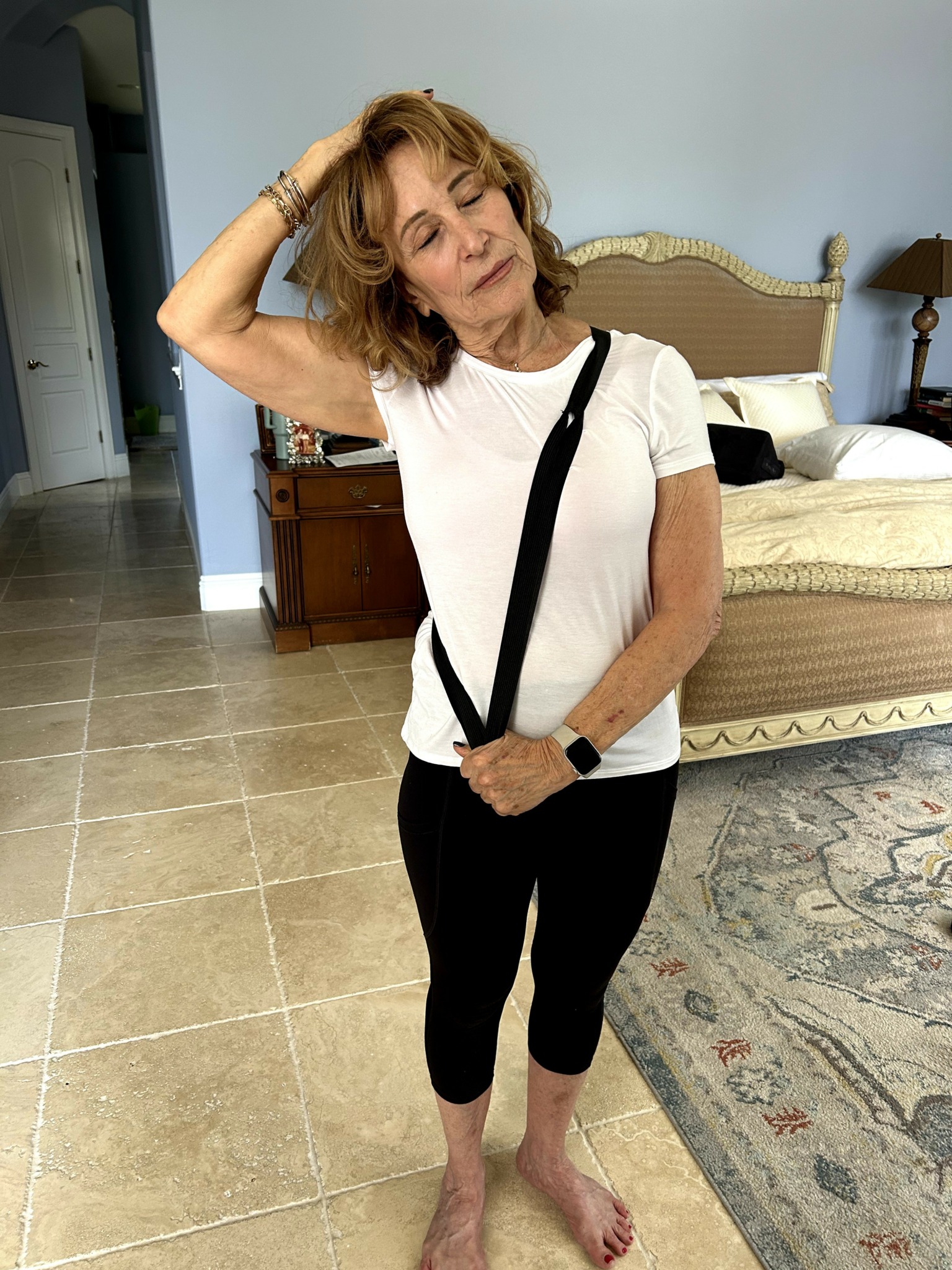 Woman with short curly hair wearing a white t-shirt and black pants, standing in a bedroom with a bed and rug.