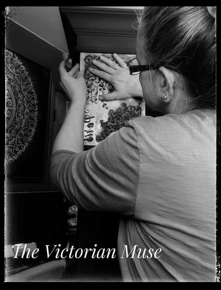 Woman with glasses and a ponytail looks at a book or artwork on a table, viewed from above.