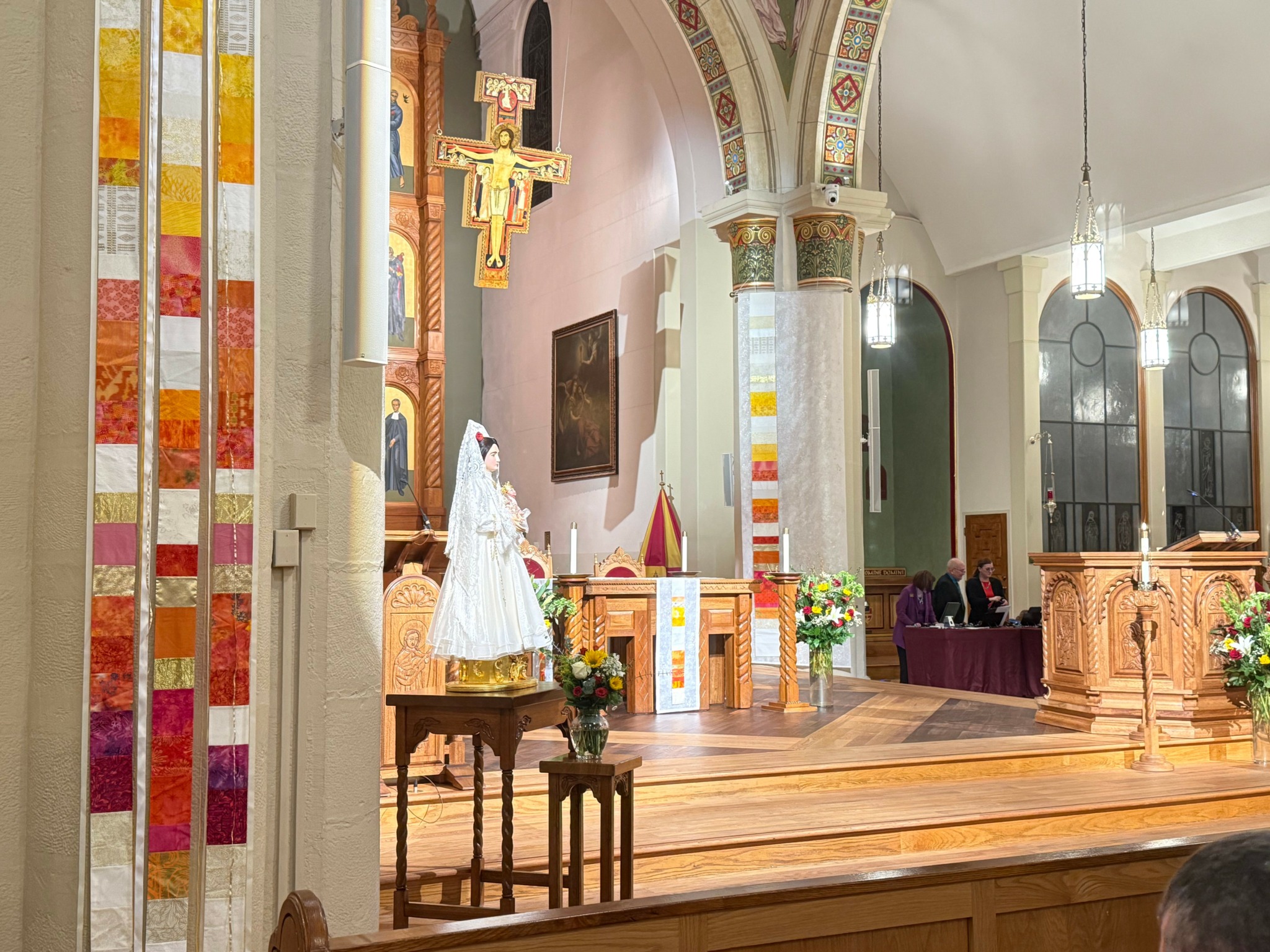 Interior of a church with altar, religious statues, and stained glass windows, decorated with flowers and lit by hanging lights.