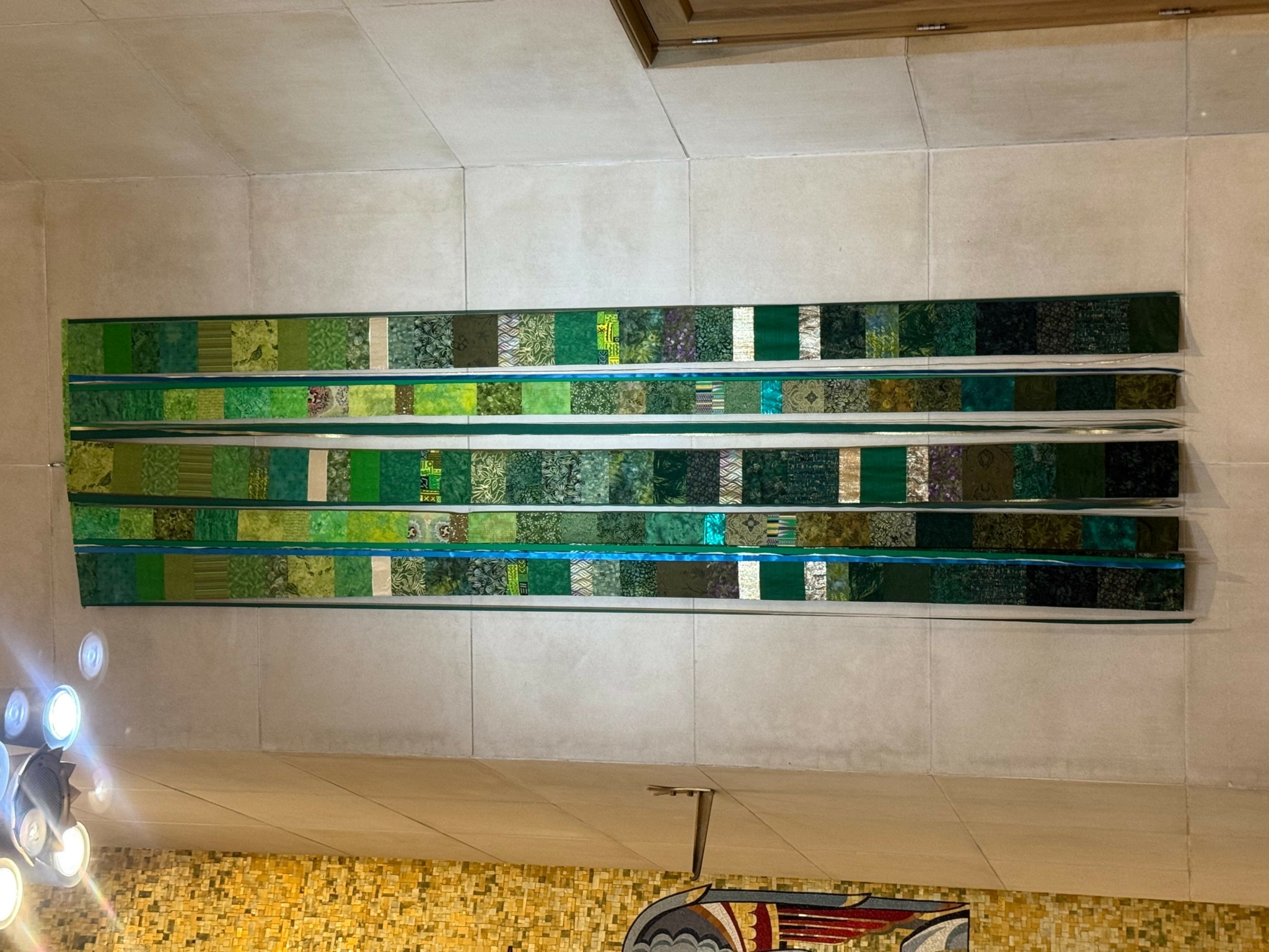 Horizontal mosaic artwork with various shades of green, blue, and beige tiles on a wall.