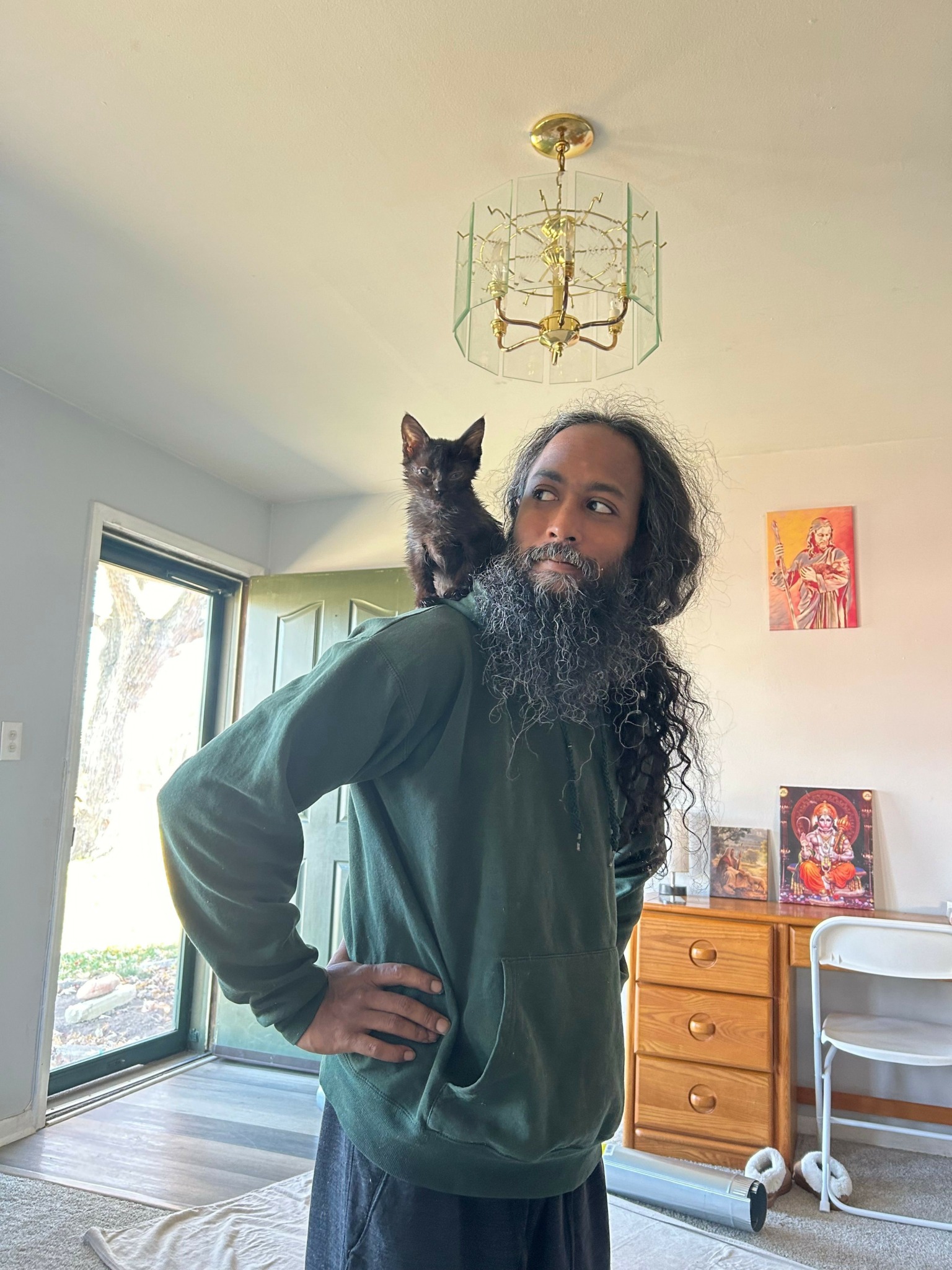 Man with long hair and beard standing indoors with a cat on his shoulder, near a window and wooden furniture.