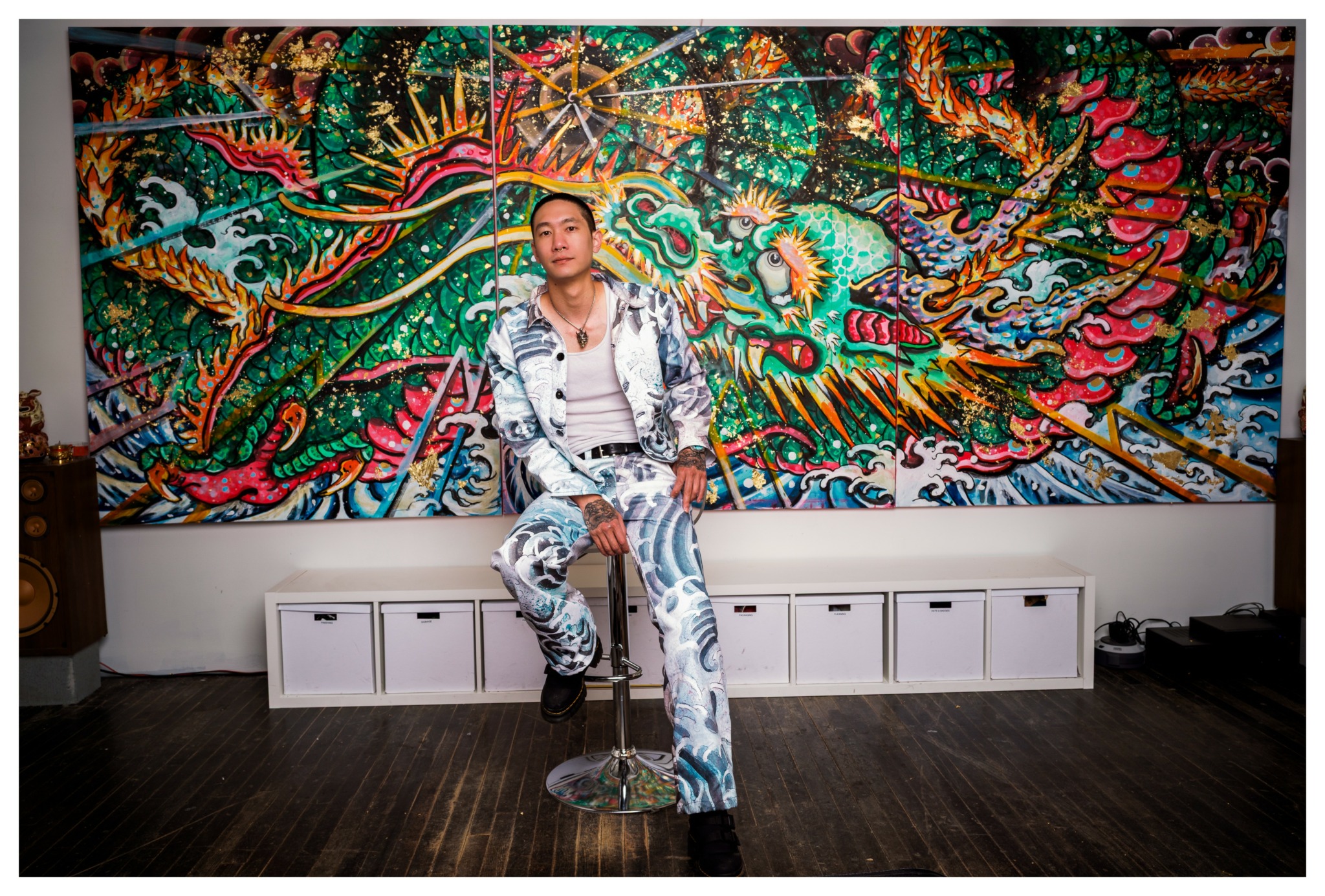 Person sitting on a stool in front of a colorful dragon mural, wearing a patterned outfit and looking at the camera.