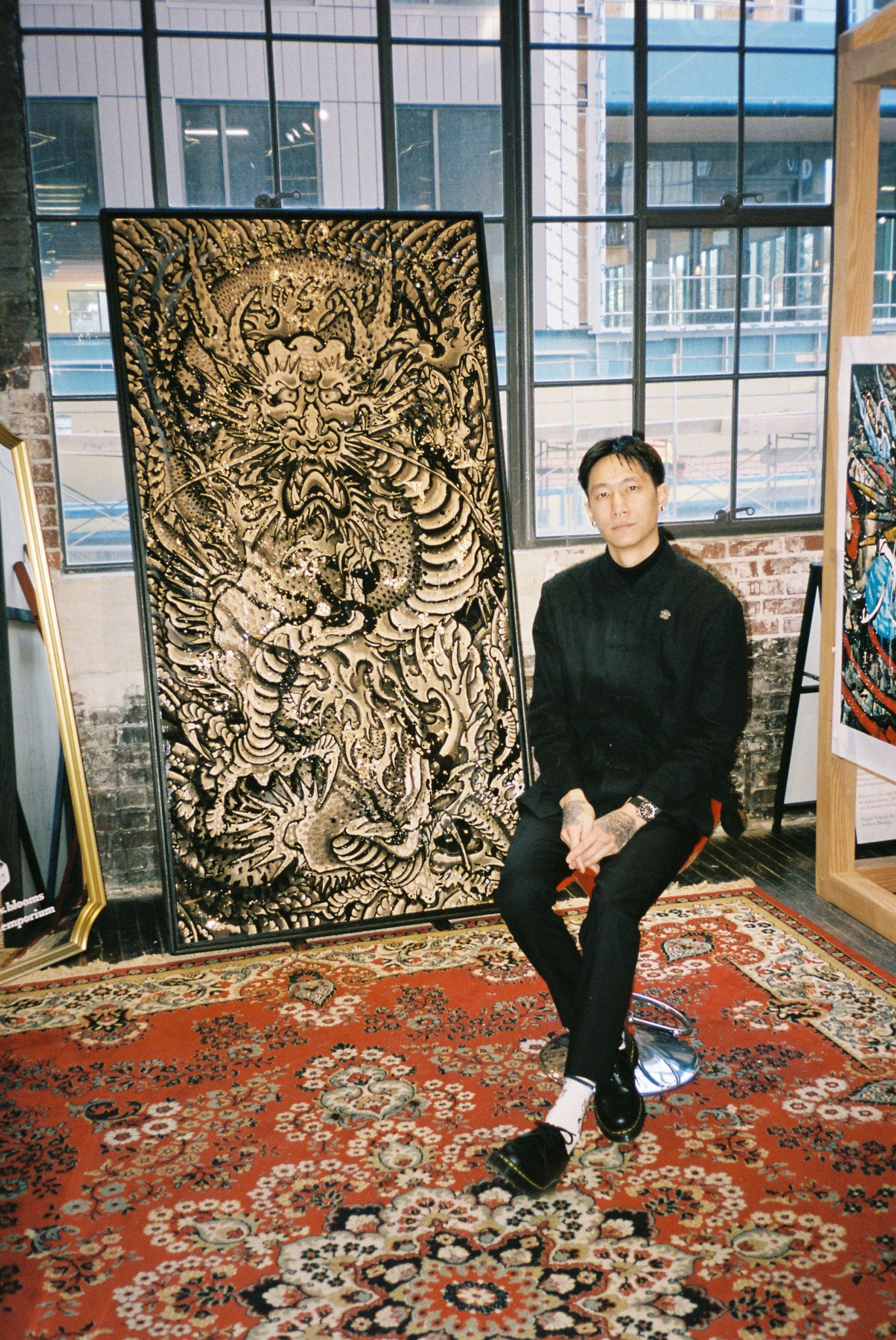 Young man sitting on a stool next to a large dragon artwork in a modern indoor space with windows and a patterned rug.